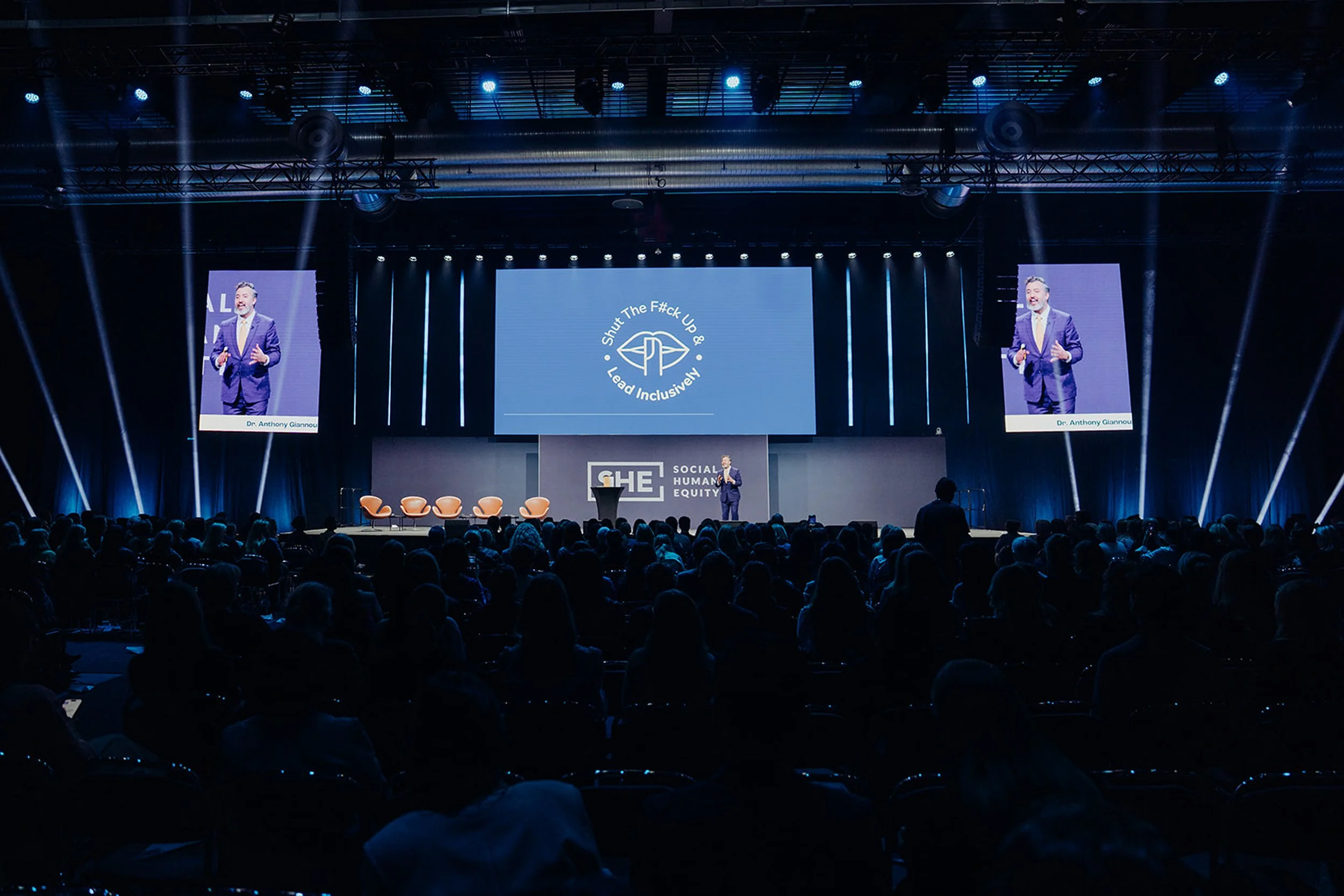 A large conference room with a stage and two large screens displaying a male speaker in a suit. The stage has a banner with the text 'GHE' and 'Social Humanity & Equity'. The main screen shows a message that says 'Shut The F#ck Up & Lead Inclusively'. The audience is seated in darkness, facing the stage.