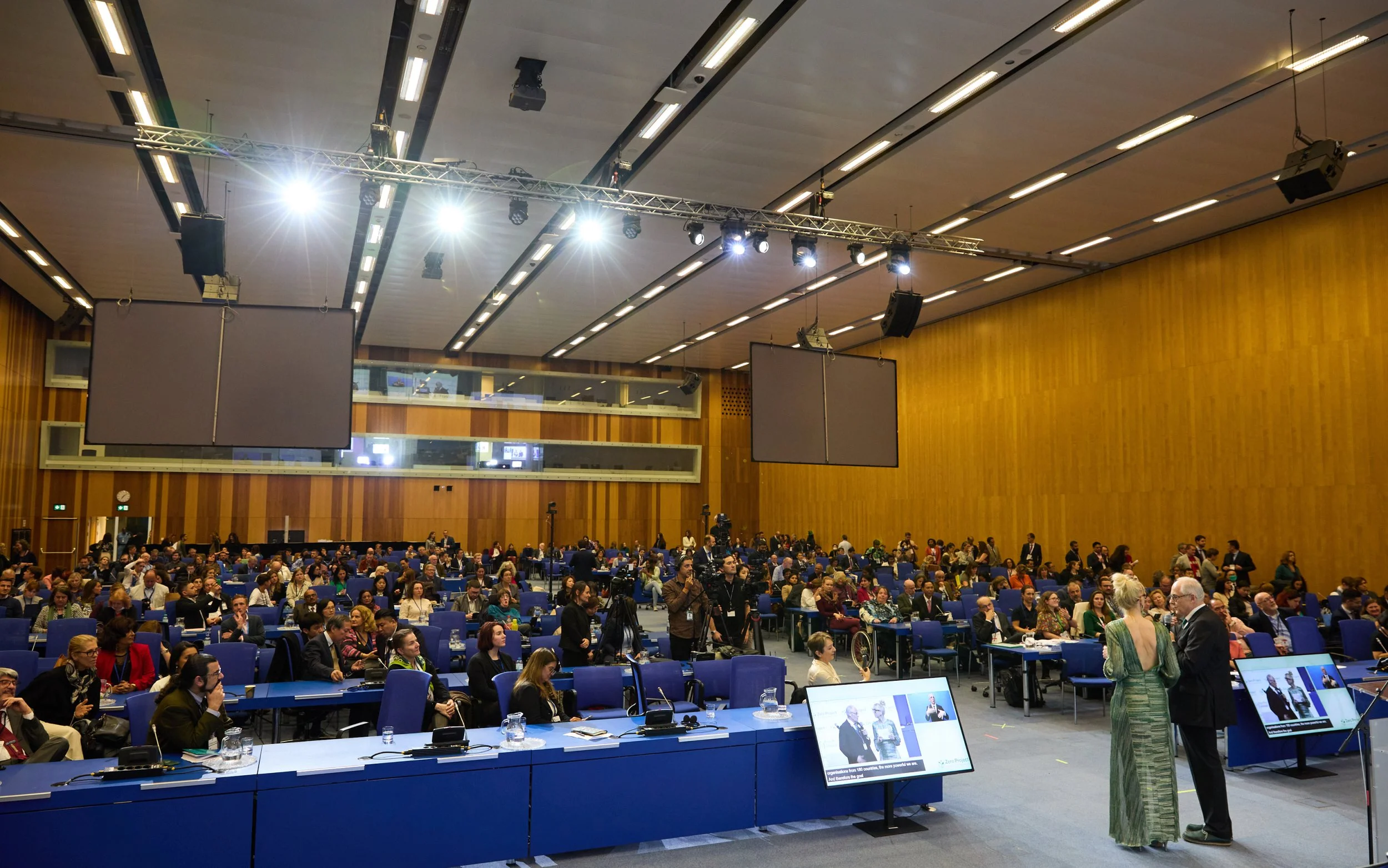 A large conference hall filled with an engaged audience, as a speaker addresses attendees from the stage during a professional event.