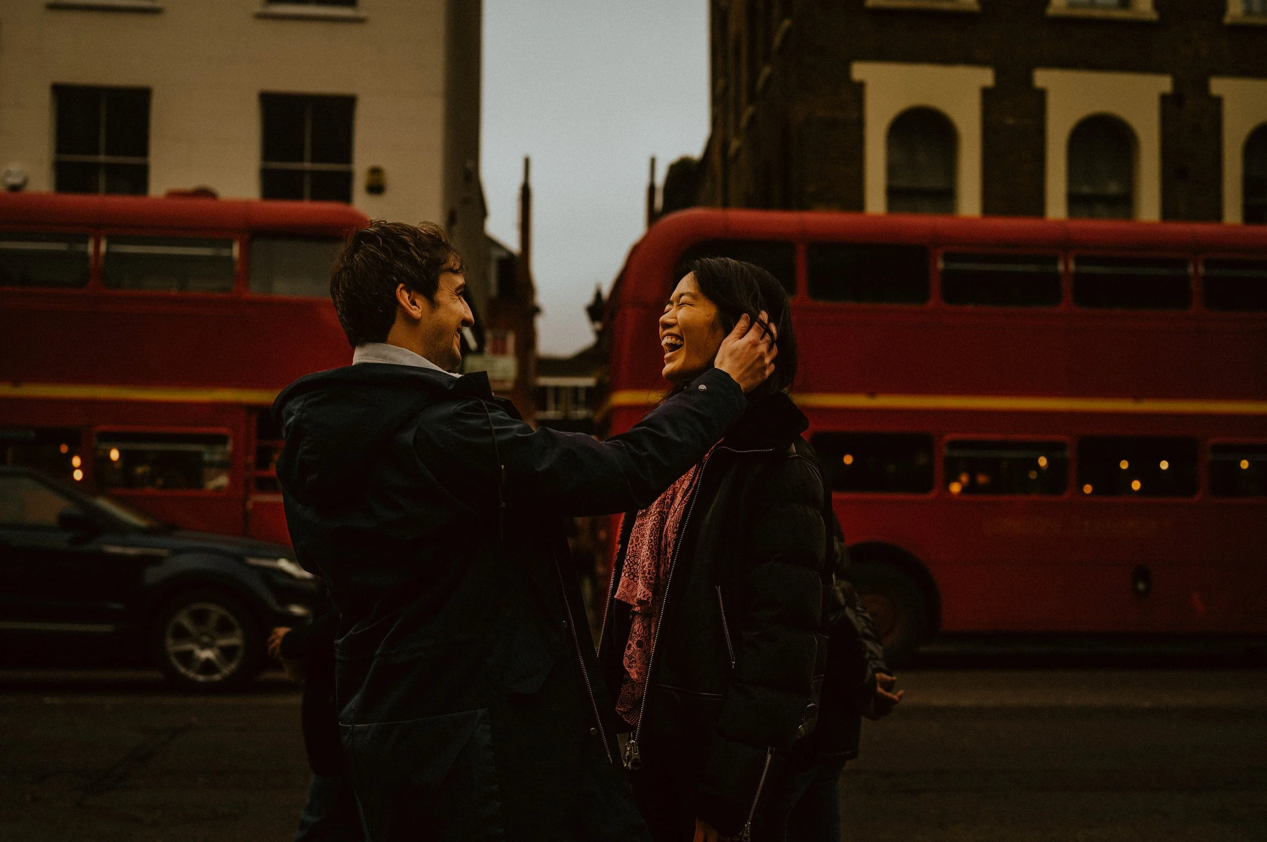 A man and a woman are smiling and laughing on a city street with a red double-decker bus in the background during the evening or early night.