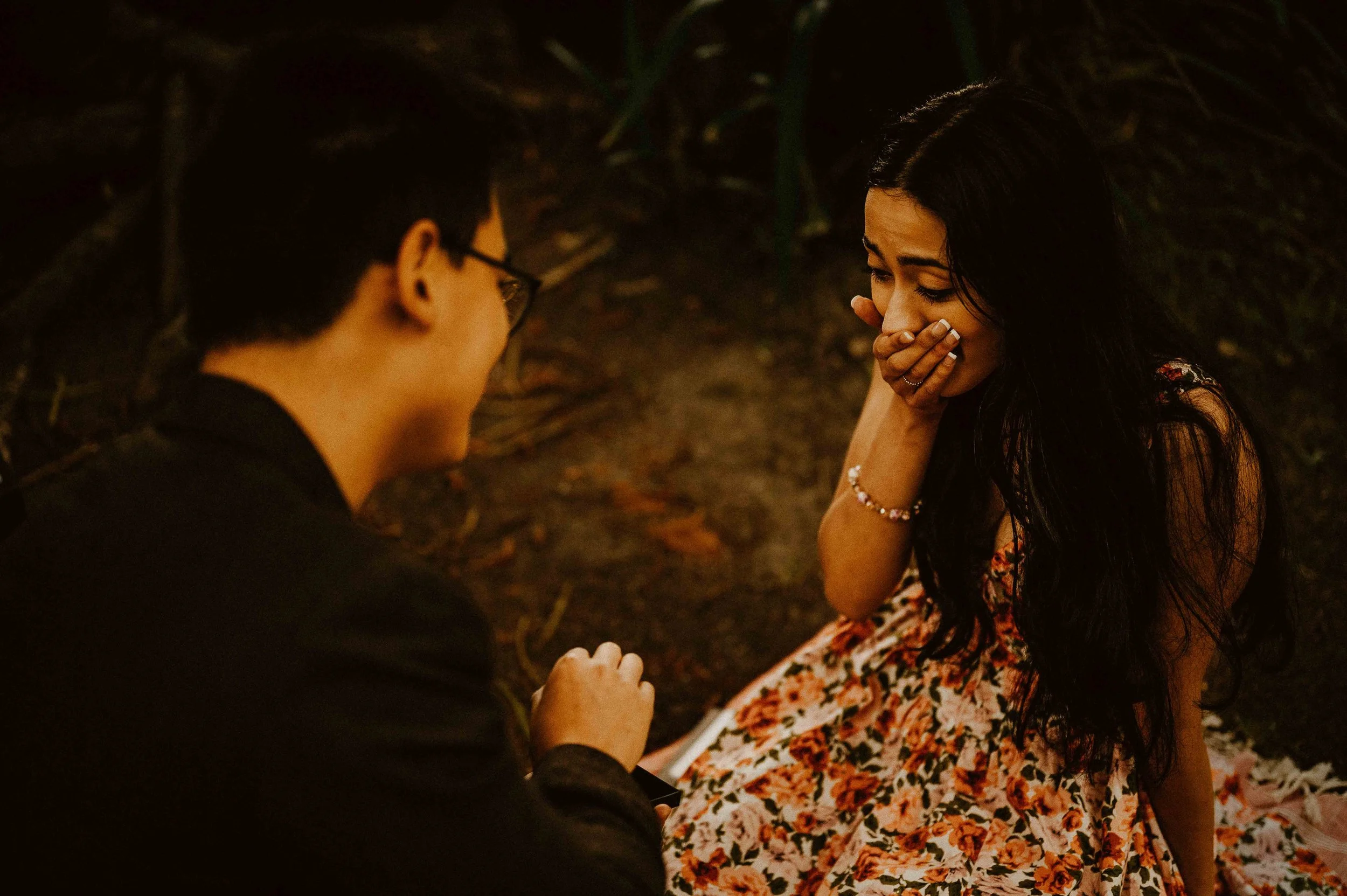 A young man kneeling in front of a young woman at night, holding her hand and proposing marriage. The woman is smiling, covering her mouth with her hand, and wearing a floral dress.