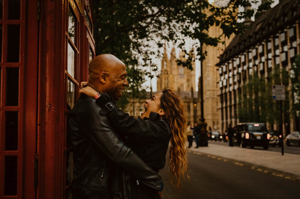 A man and woman are playfully hugging and laughing on a city street at sunset. The man is bald and dressed in a black leather jacket, leaning against a red telephone booth. The woman has long, wavy hair and is also wearing a black jacket. There are cars and tall buildings in the background.