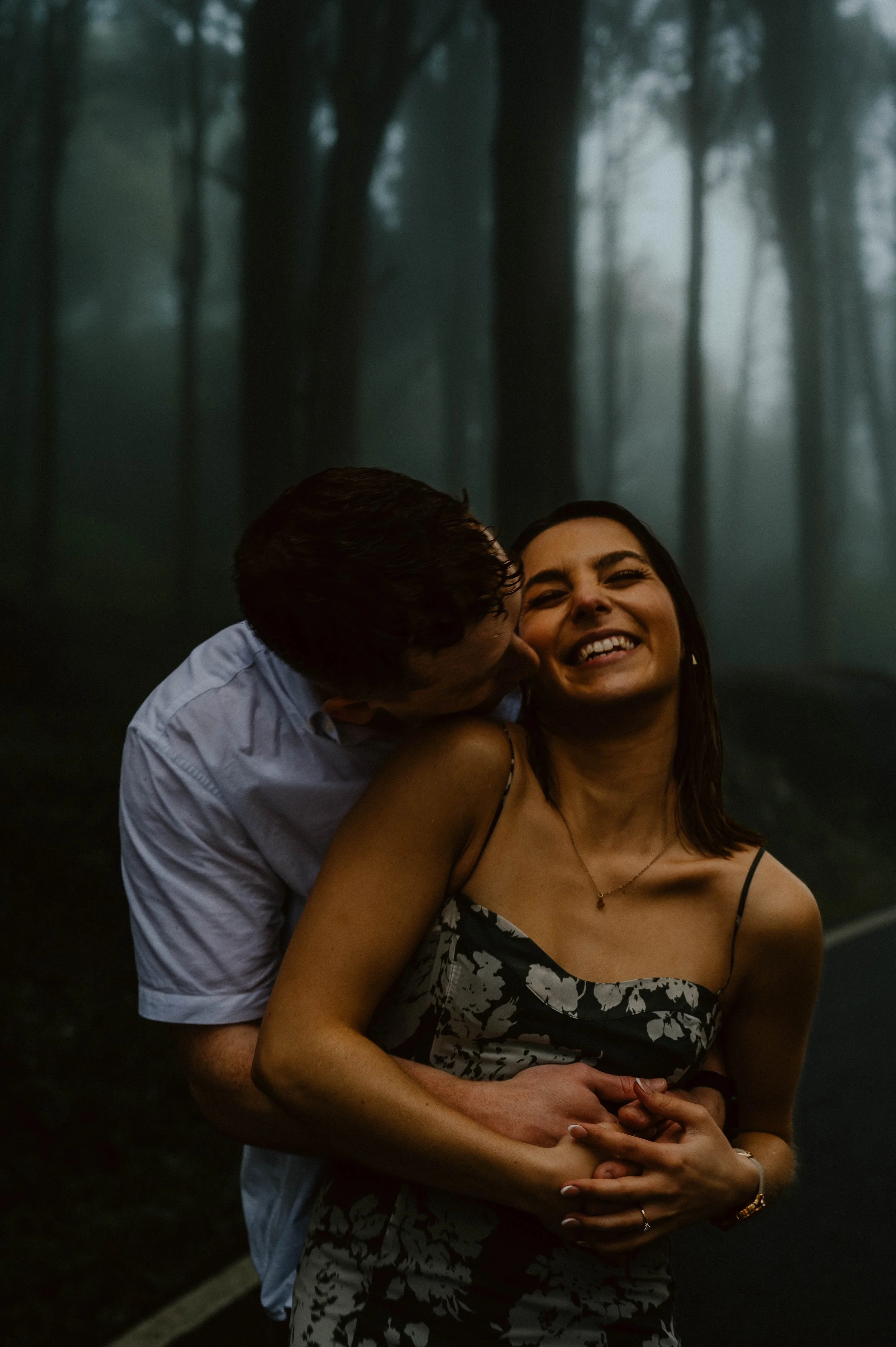 A couple embracing outdoors in a misty forest. The man is kissing the woman on the cheek, and she is smiling with closed eyes, wearing a floral dress.