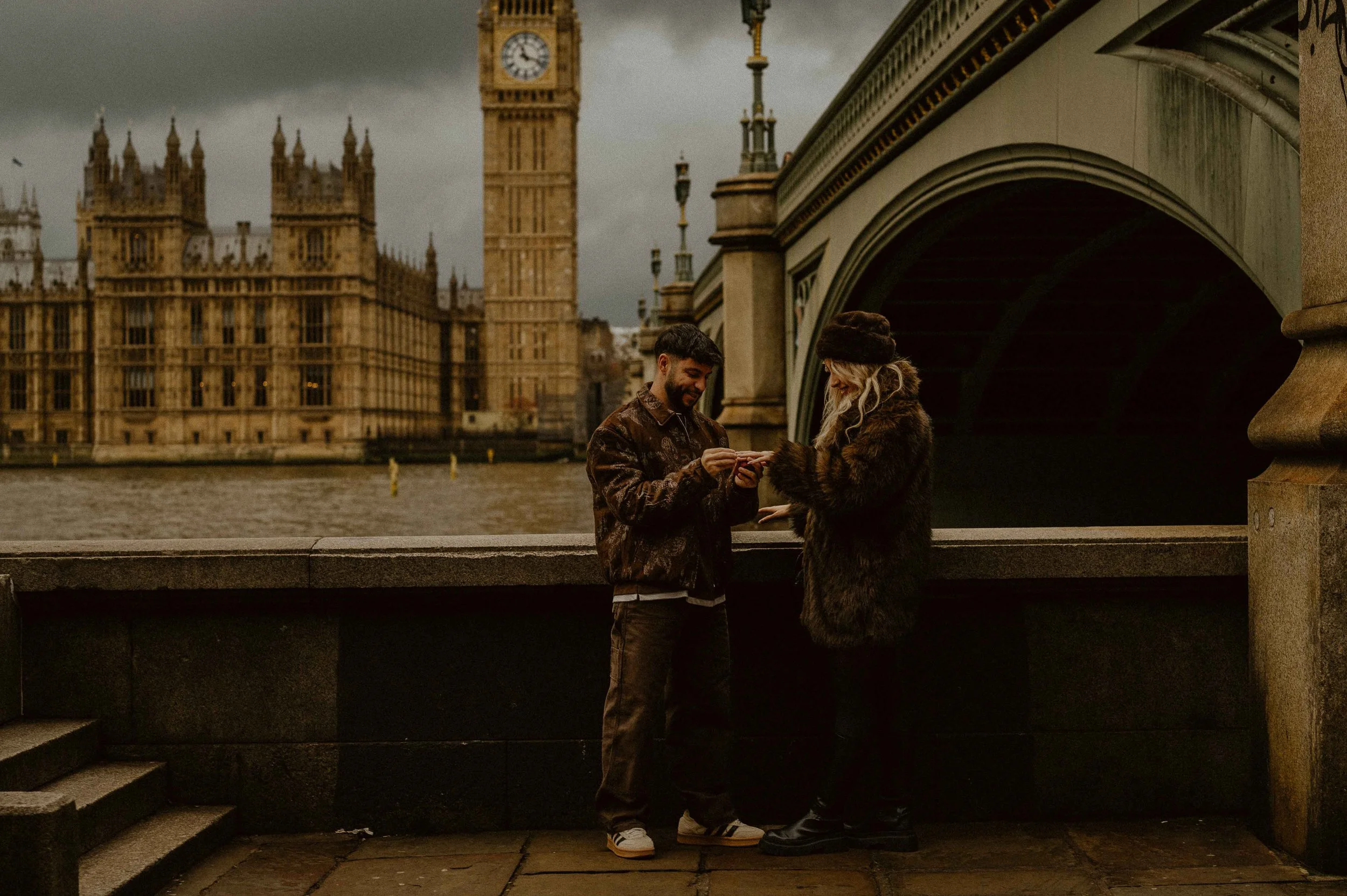 Two people standing by the riverbank in front of a bridge, exchanging rings, with the Big Ben clock tower and the Houses of Parliament in the background in London, under a cloudy sky.
