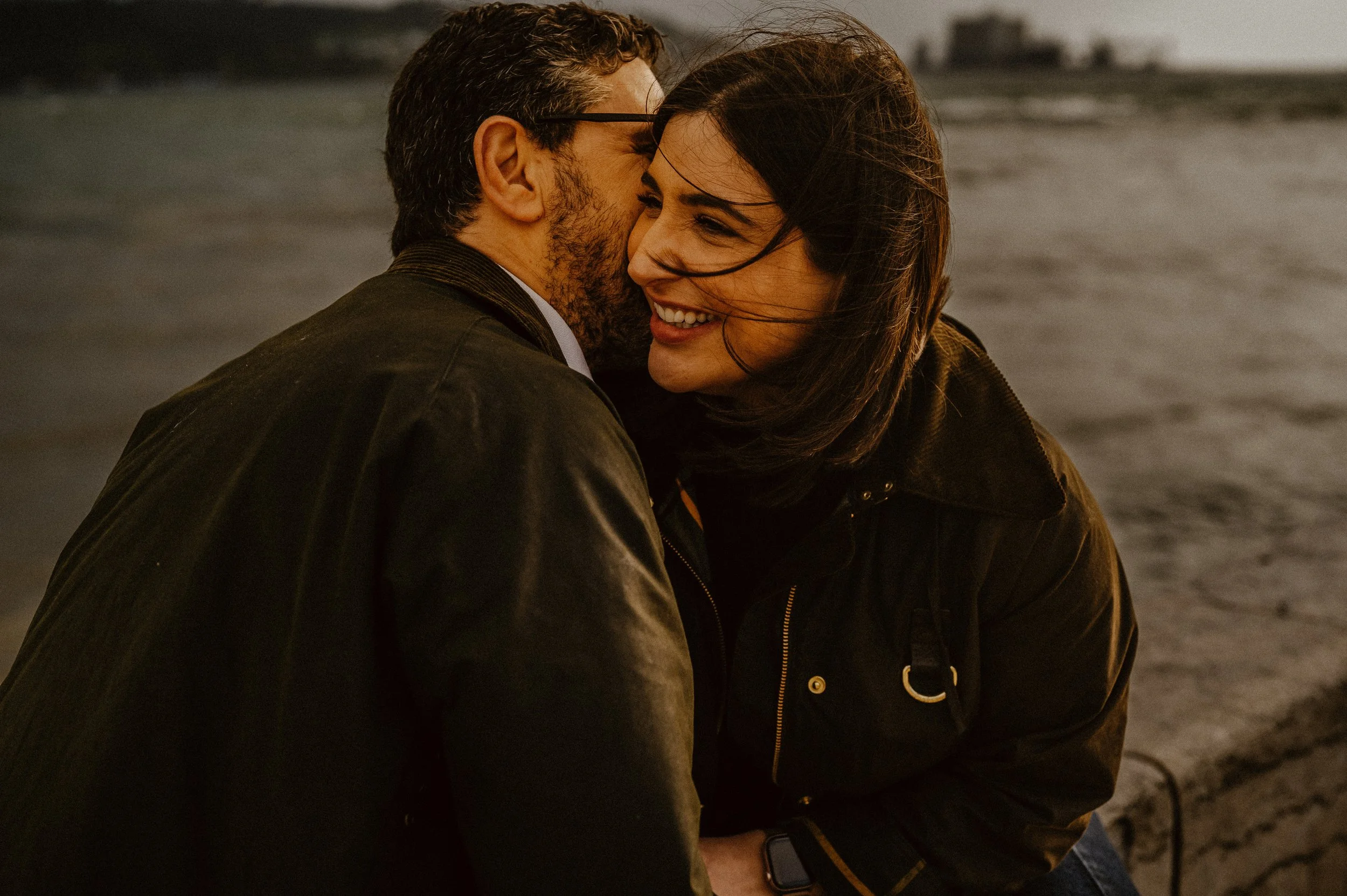 A man and woman smile and lean close together on a beach during sunset or dusk, with waves in the background.