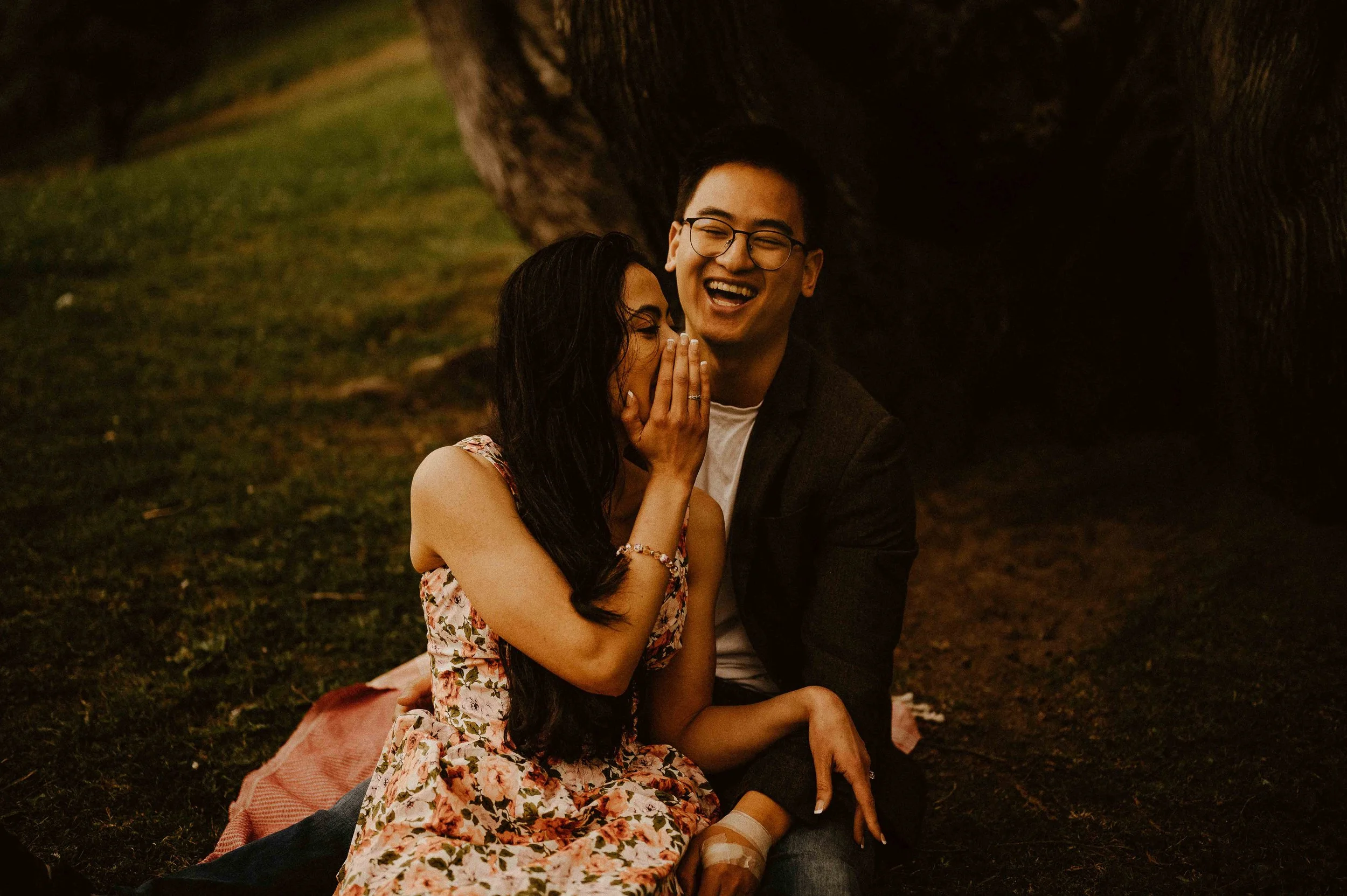 A young woman and a young man sit close together on the ground outside at night, sharing a joyful moment. The woman, with long dark hair and wearing a floral dress, covers her mouth as the man laughs, wearing glasses and a dark jacket. They are sitting on a blanket under a large tree, with grass around them and a dark background.