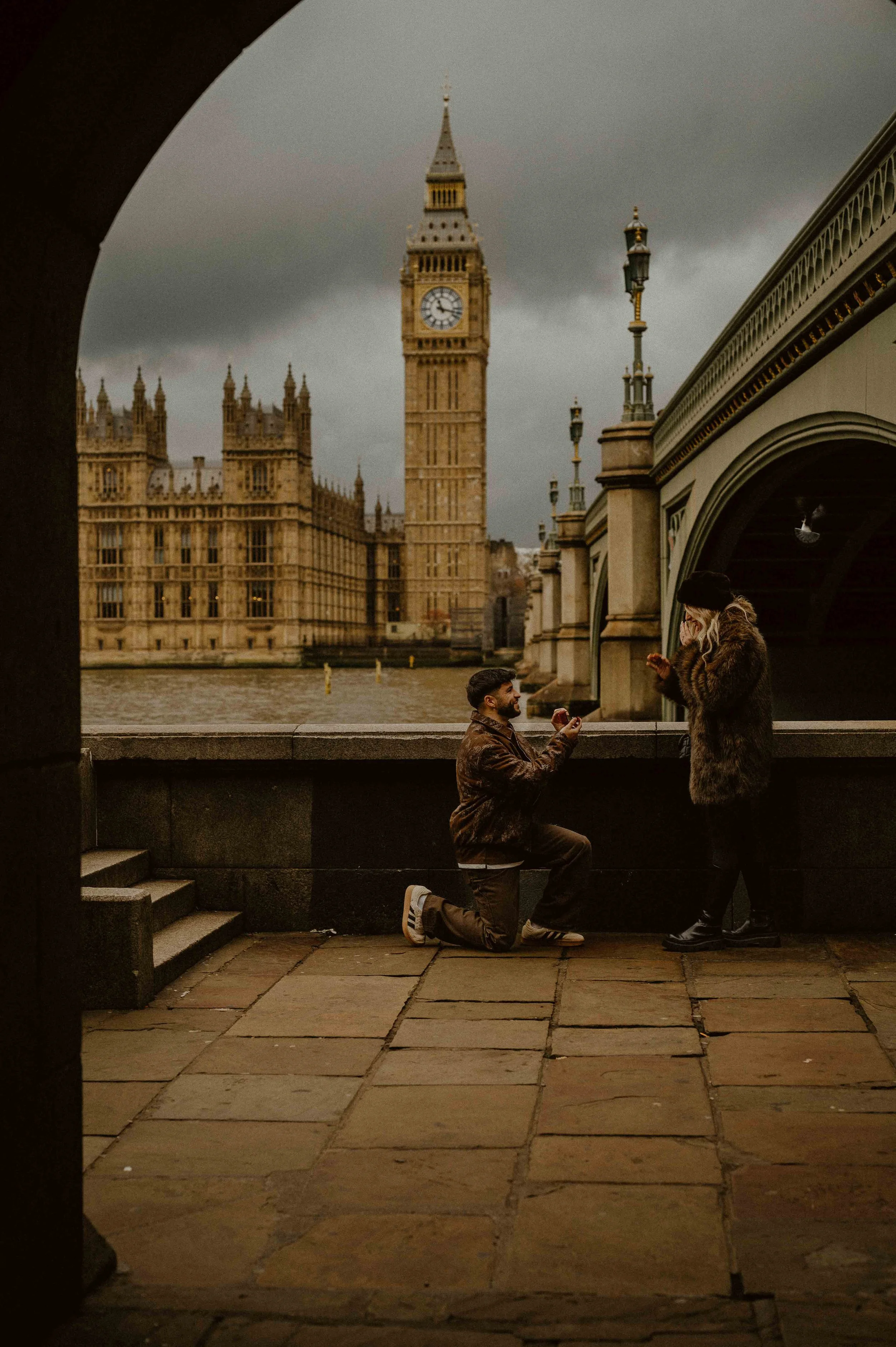 A man kneeling on one knee proposing to a woman on a bridge with Big Ben in London in the background.