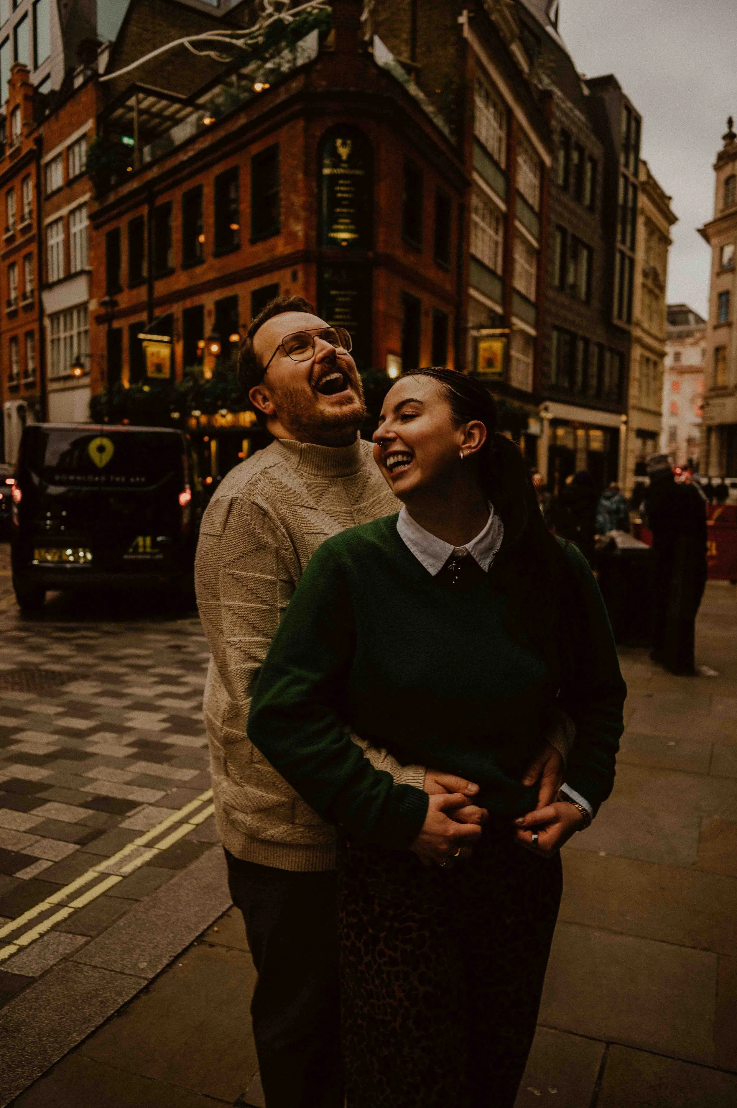 London couples photographer and her couple, right before their proposal, in Soho. The man hugs the woman from behind and they both laugh in this connection-focused experience in London