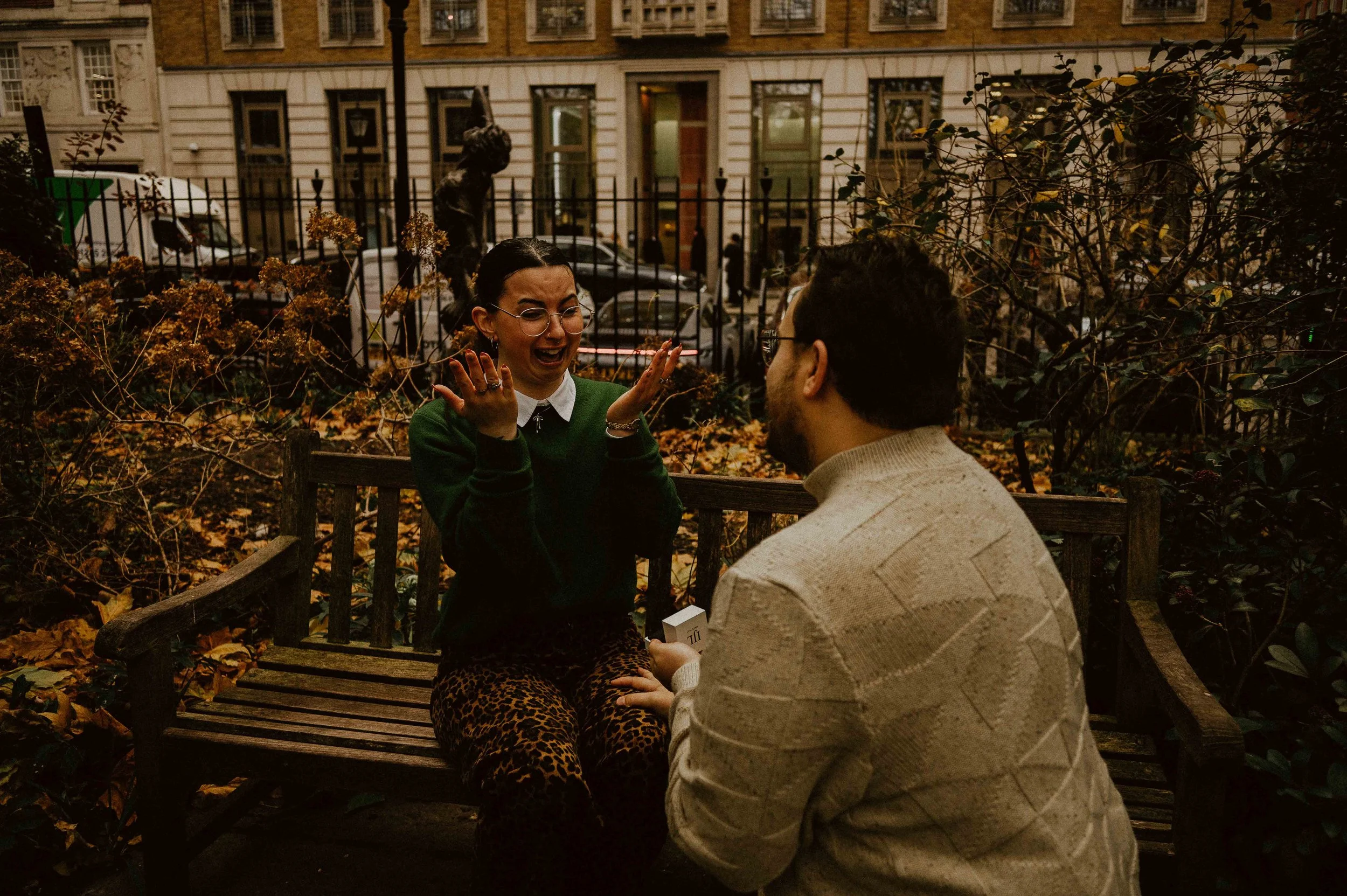 A woman with glasses and dark hair laughing and gesturing with her hands, sitting on a park bench across from a man with glasses and a beard, in an urban park surrounded by autumn leaves and bushes, with a building and parked cars in the background.