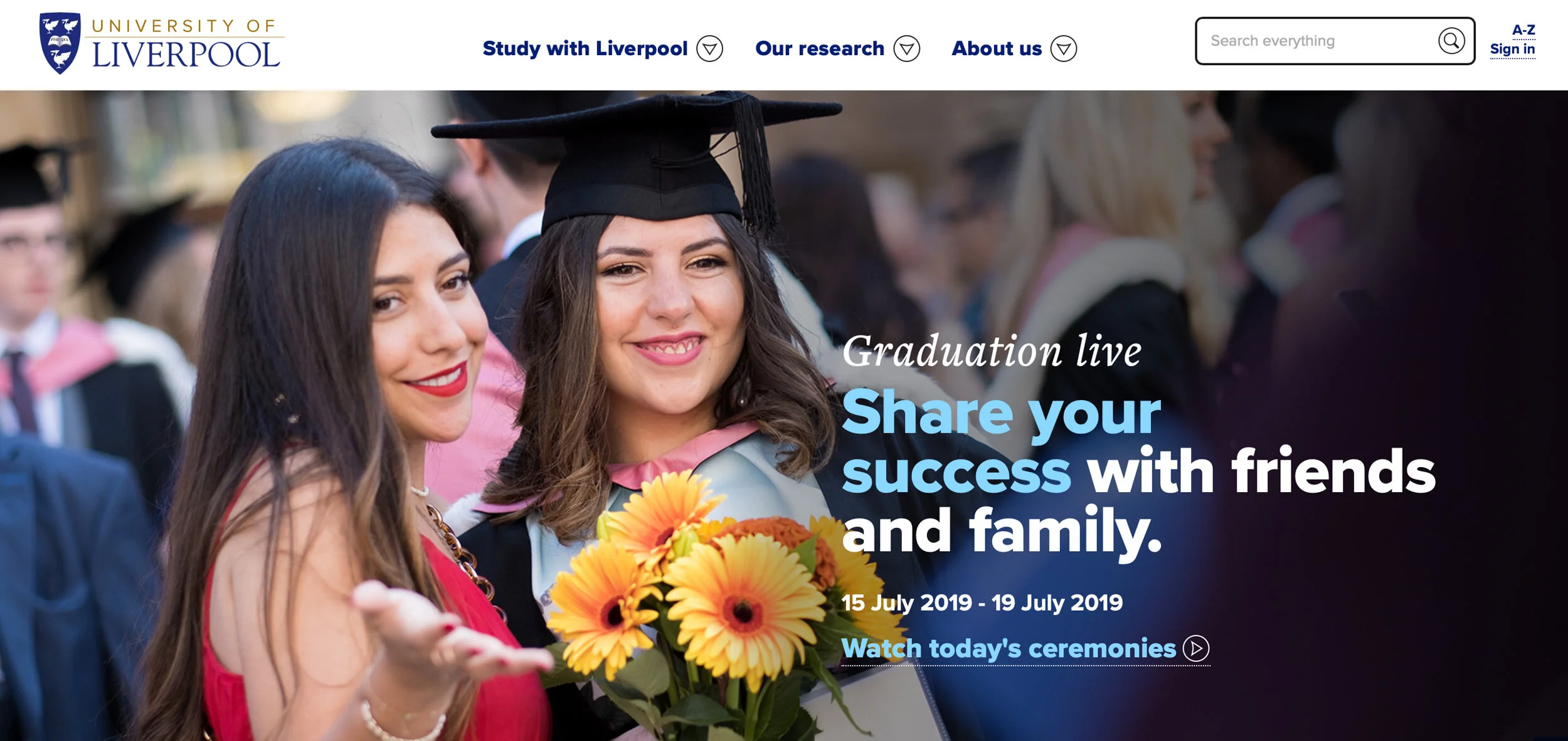 Photograph of two female graduates smiling and celebrating at a university graduation ceremony, one holding a bouquet of yellow flowers.
