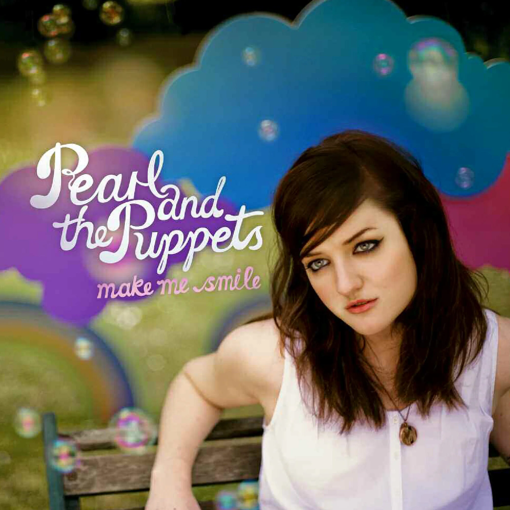 Album cover for Pearl and the Puppets titled "Make Me Smile" showing a woman with dark brown hair and eyeliner, sitting on a park bench with colorful bubbles and background scenery.