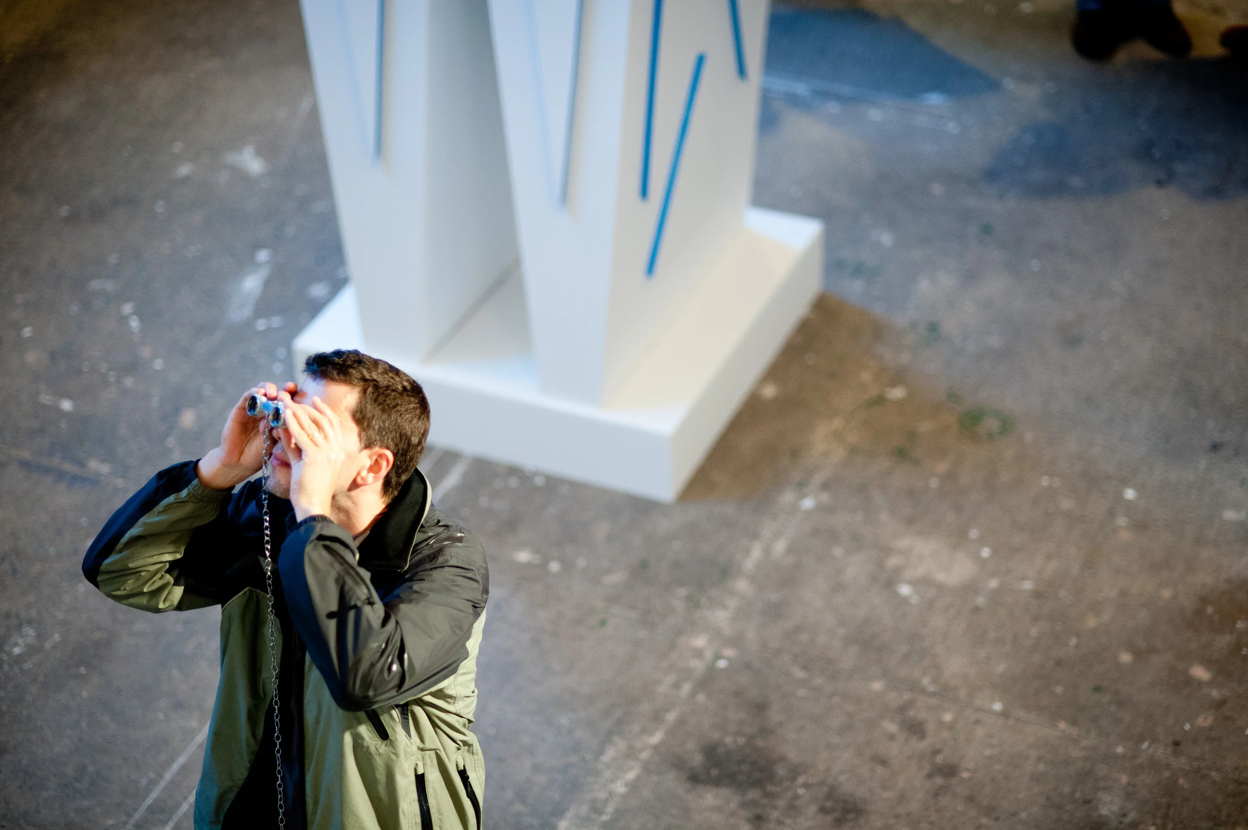 A man wearing a black and green jacket looking through binoculars on a concrete floor with a white abstract sculpture in the background.