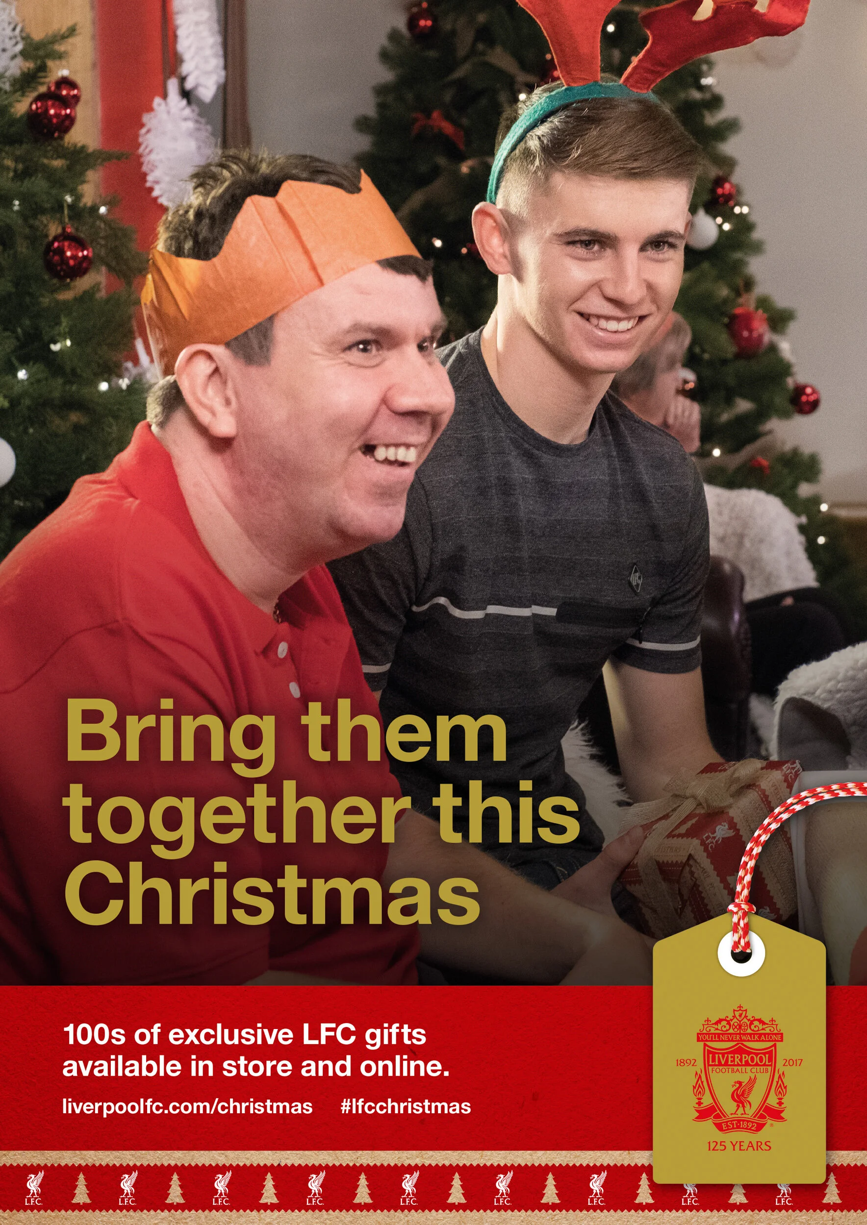 Two smiling men sitting in front of Christmas trees during a festive celebration, wearing reindeer antlers and a paper crown, exchanging gifts.