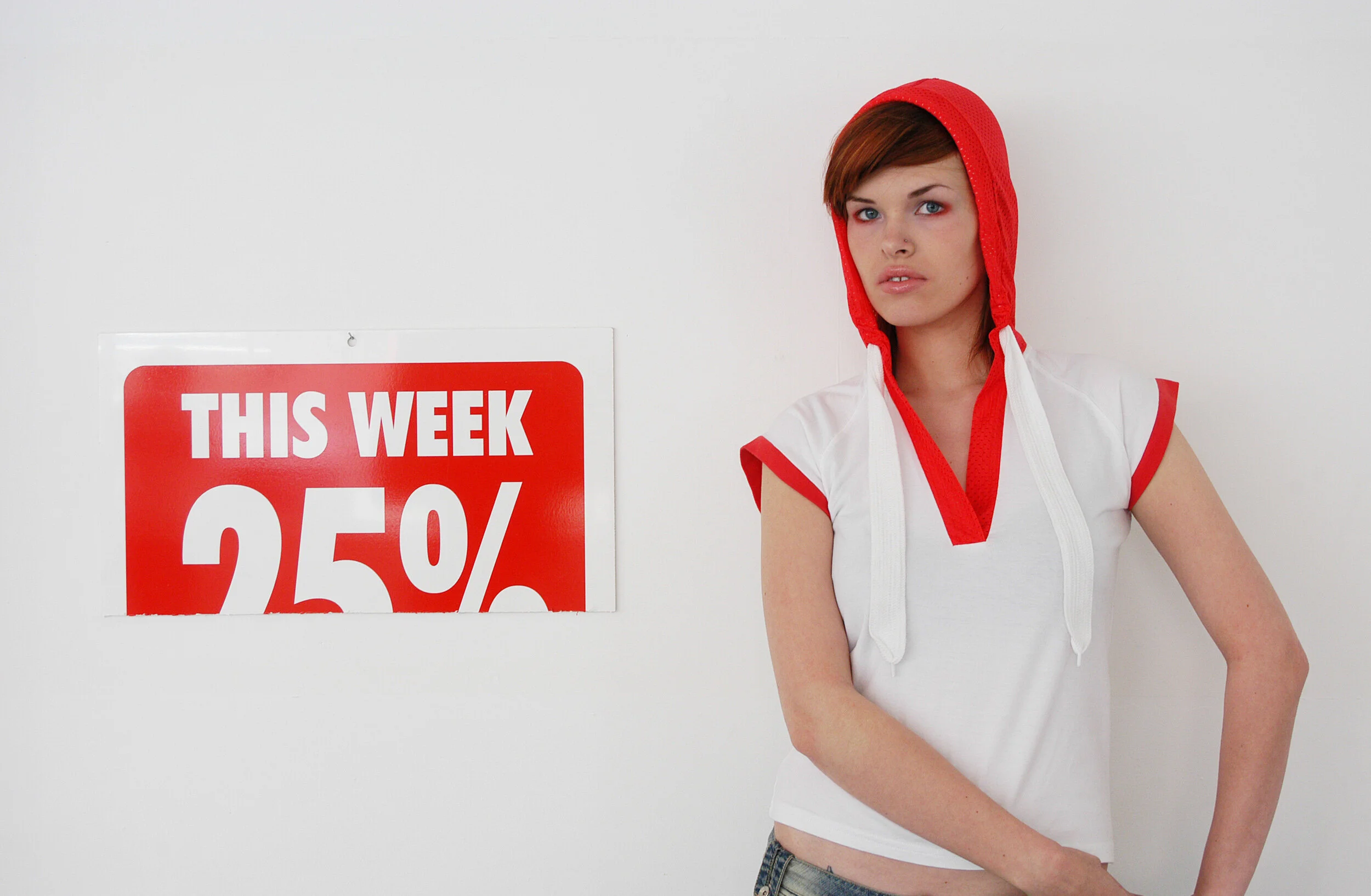 Young woman with red hair wearing a white top with red trim and a red hood, standing next to a white wall with a red and white sign that indicates a 25% discount for the week.