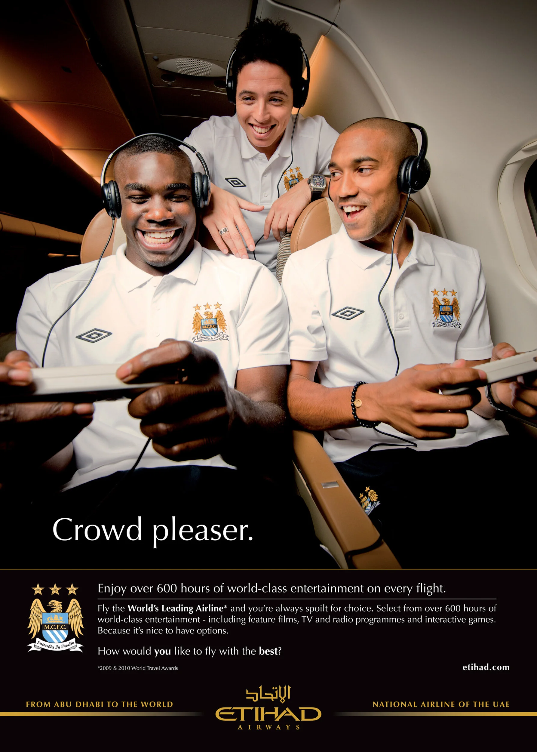 Four airline crew members, two men sitting in the aircraft, and two women standing behind them, smiling and looking at a device. They are wearing white uniforms with a badge, and the scene appears to be inside an airplane.