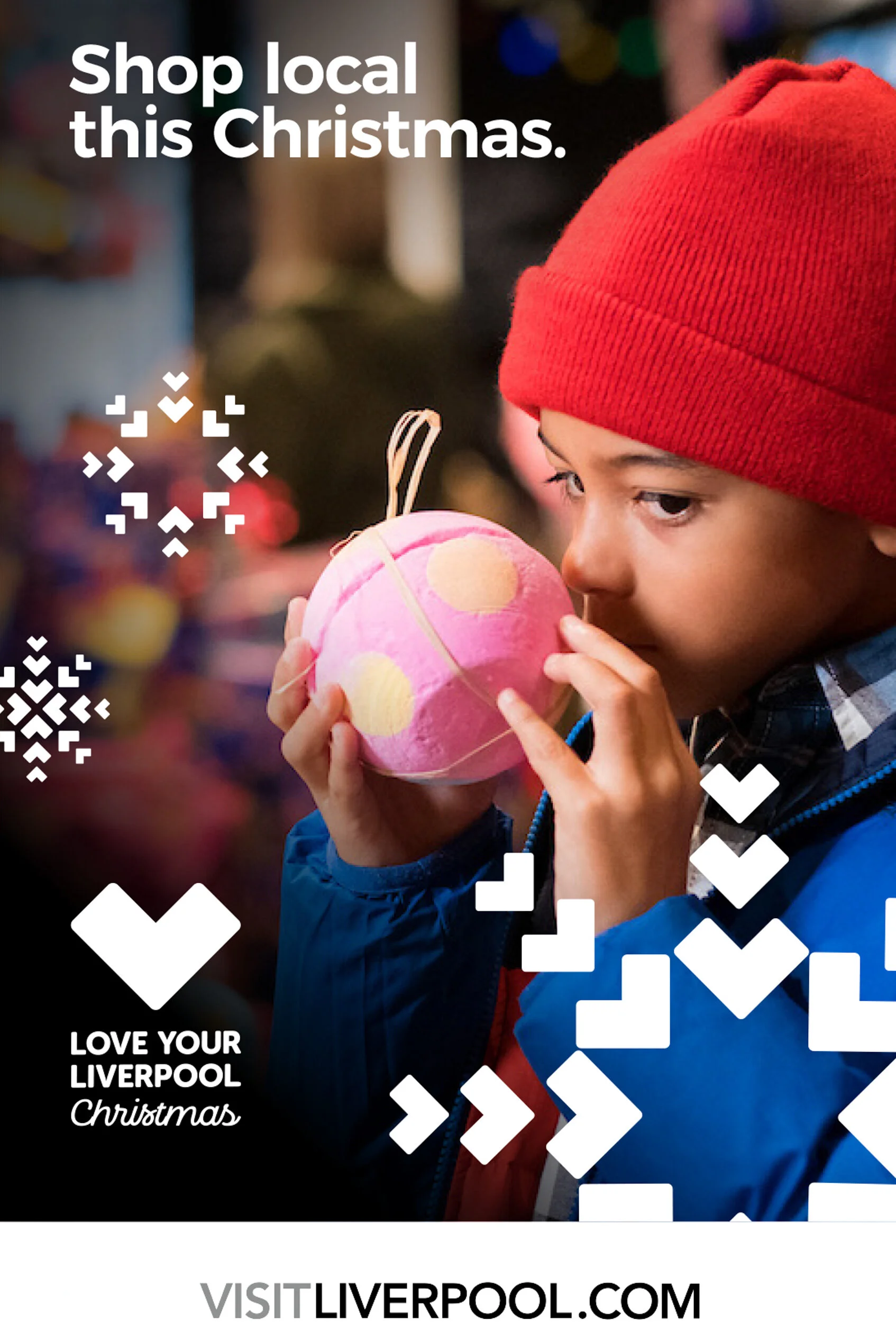 A young boy dressed in a blue jacket and red beanie is smelling a pink and yellow polka-dotted Christmas ornament.