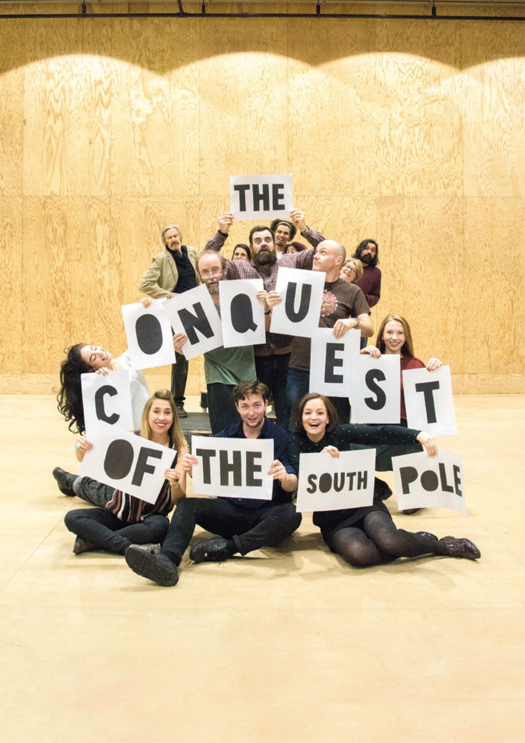 Group of people holding signs that spell out "THE QUEEN'S OF THE SOUTH POLE," inside a wooden hall.