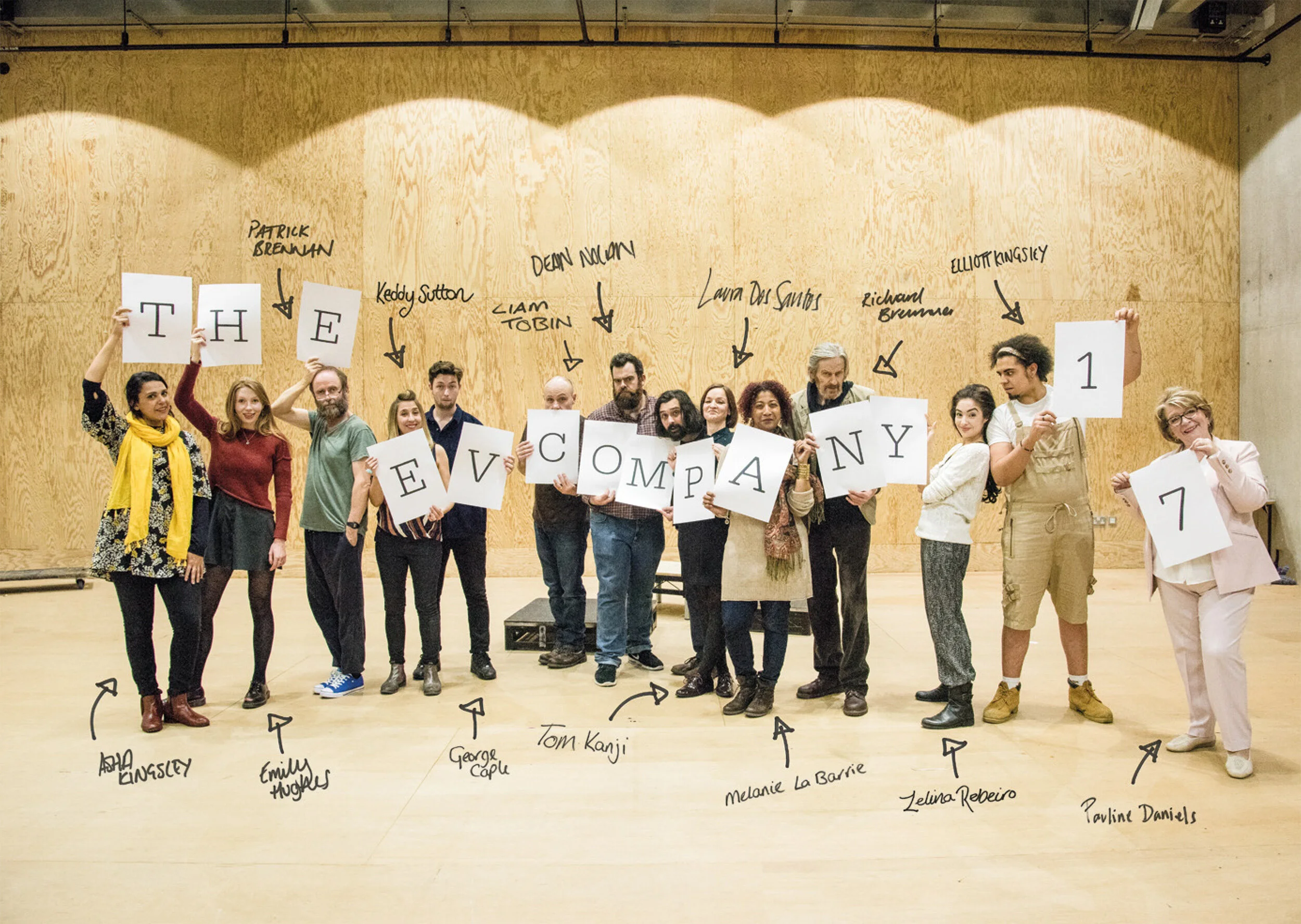 Group of people standing in a line holding large white cards with black letters spelling out "THE EV COMPANY 1 7" in front of a wooden wall with signatures and annotations.