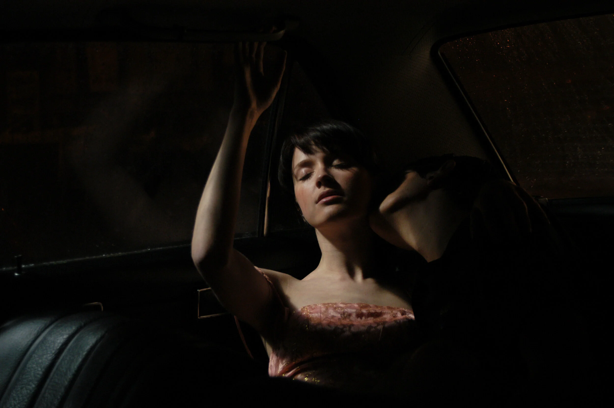 A woman with short dark hair sitting in a car at night, eyes closed, head tilted back, hand on the ceiling, wearing a strapless pink dress.