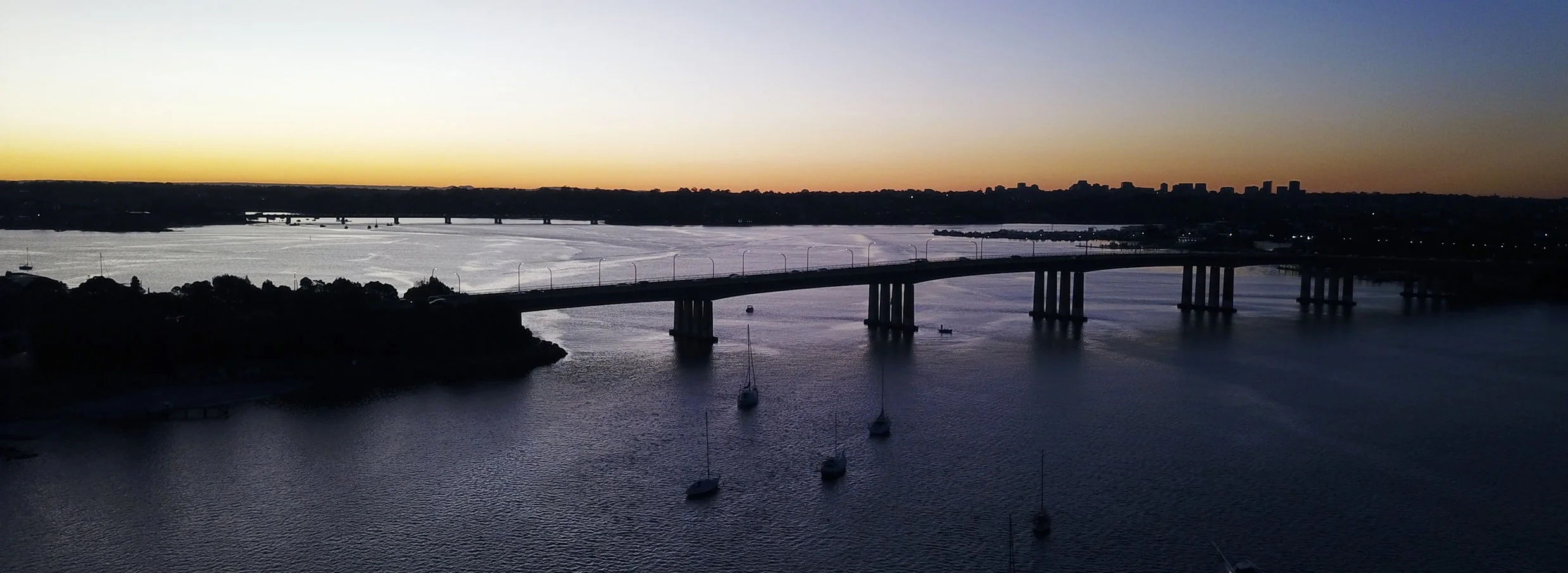 Captain Cook bridge in Sydney's south suburb the Sutherland Shire over George's River at sunset, with sailboats moored below and city skyline in the distance.