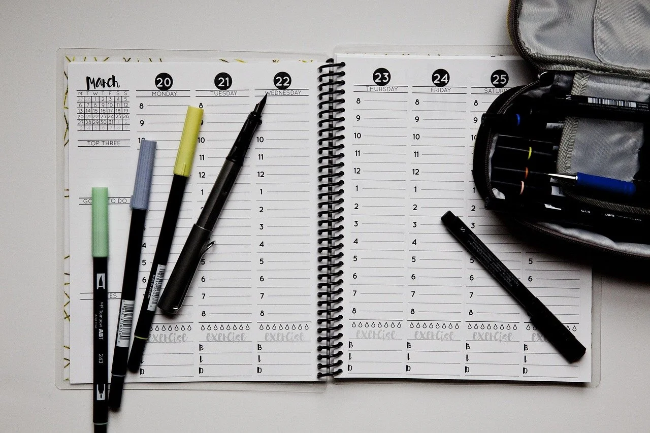 A weekly planner lying on a desk. There are fine-tipped markers with different colors on top of he notebook. It is open to a page that says March.
