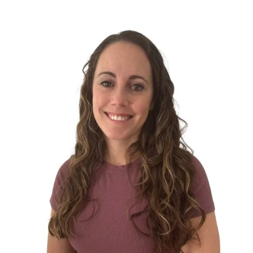 A woman with long, wavy brown hair wearing a mauve short-sleeved top, smiling, against a plain white background.