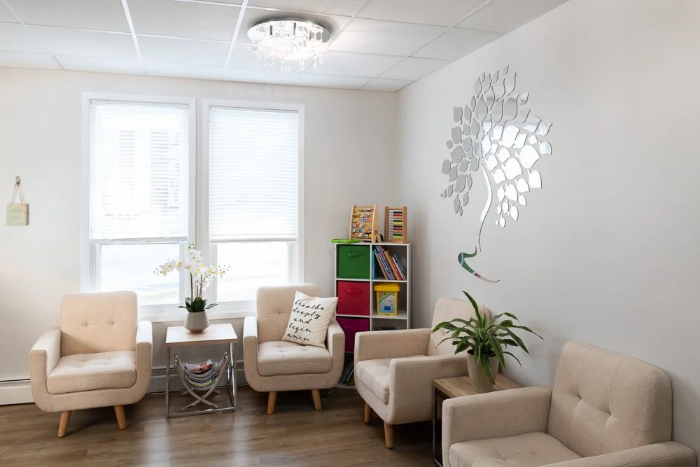 Waiting room at Spirit Psychological with cream chairs, wood floor, plants, shiny logo wall decal, and shelf with books and toys.