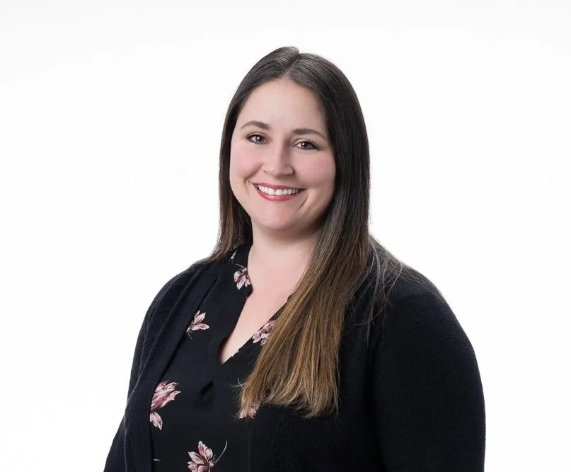 A woman with long brown hair smiling, wearing a black top with pink floral patterns and a black cardigan, against a plain white background.