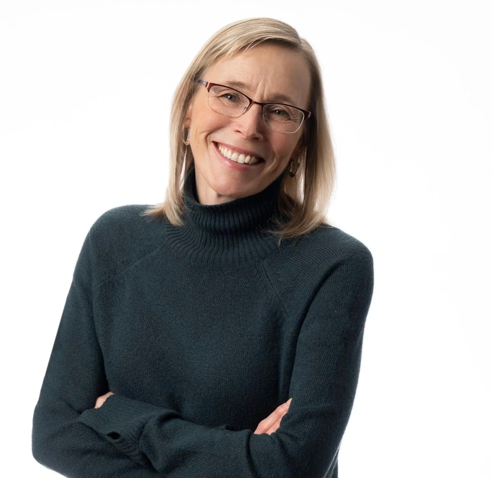 A middle-aged woman with blonde hair, glasses, and a black turtleneck sweater smiling with arms crossed against a white background.