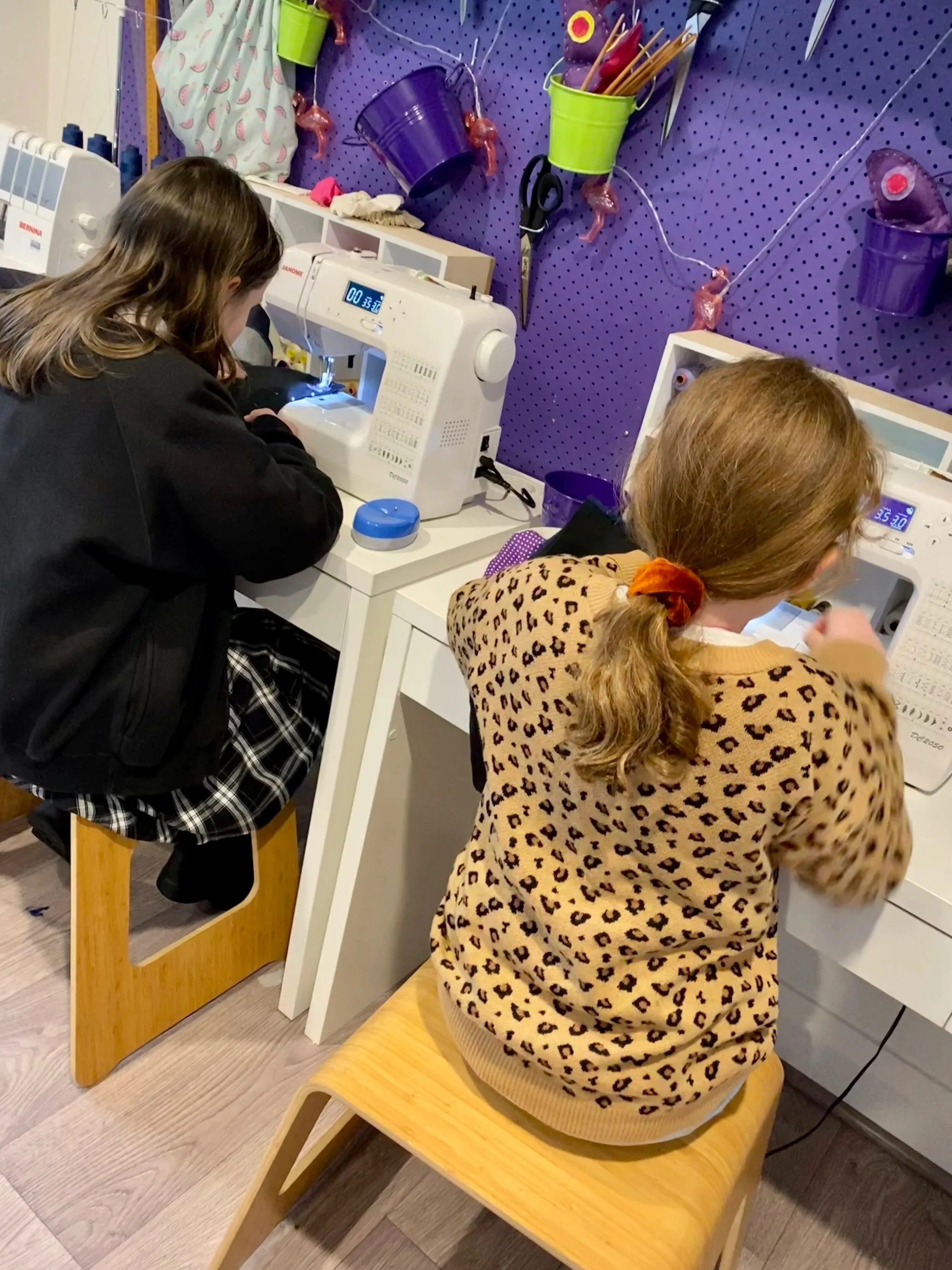 Two young girls sewing on machines at a craft table with purple wall and colorful buckets for supplies.
