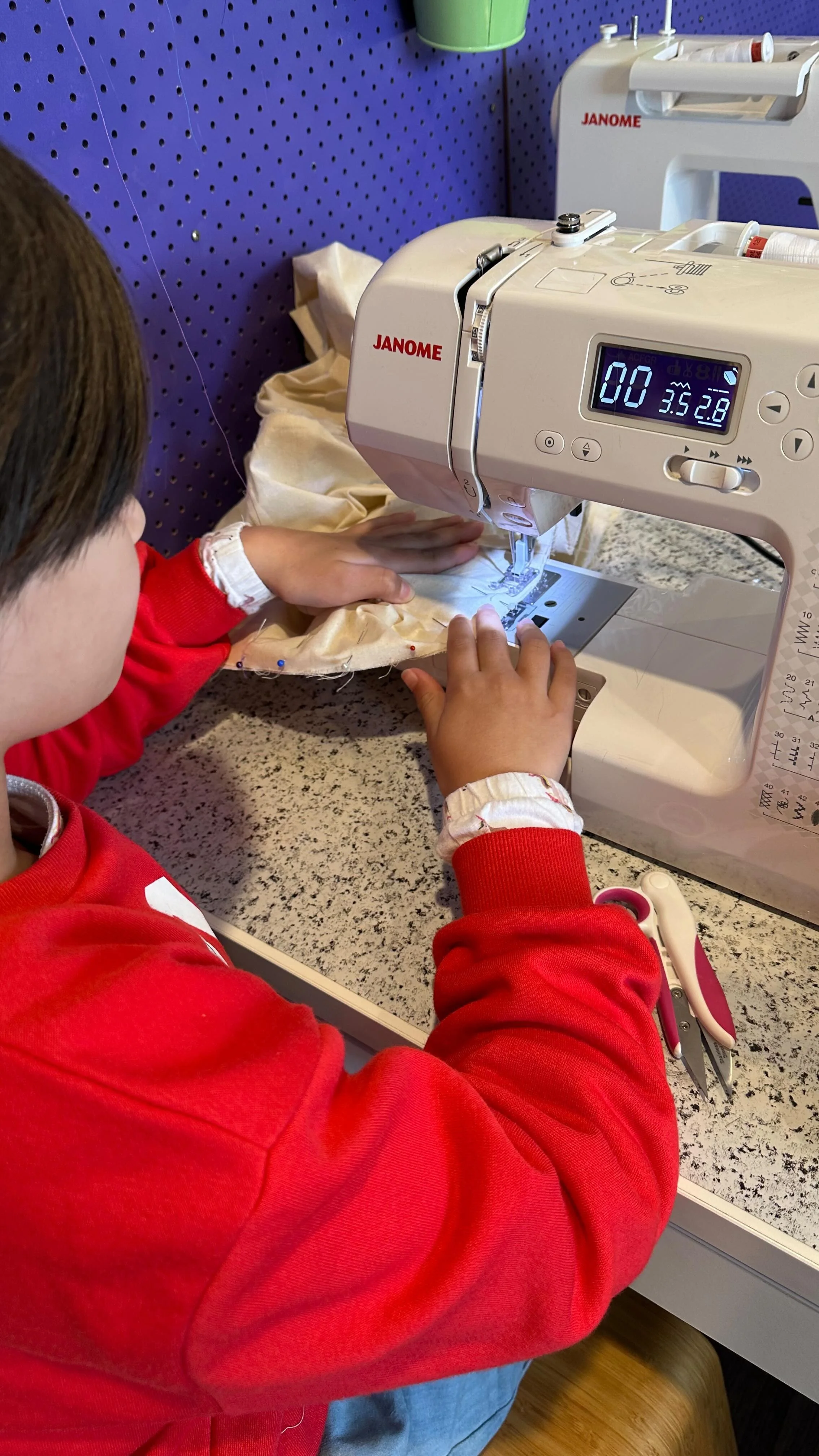 A child sewing fabric on a Janome sewing machine, sitting at a speckled countertop with scissors nearby, in a craft room with a purple pegboard wall.