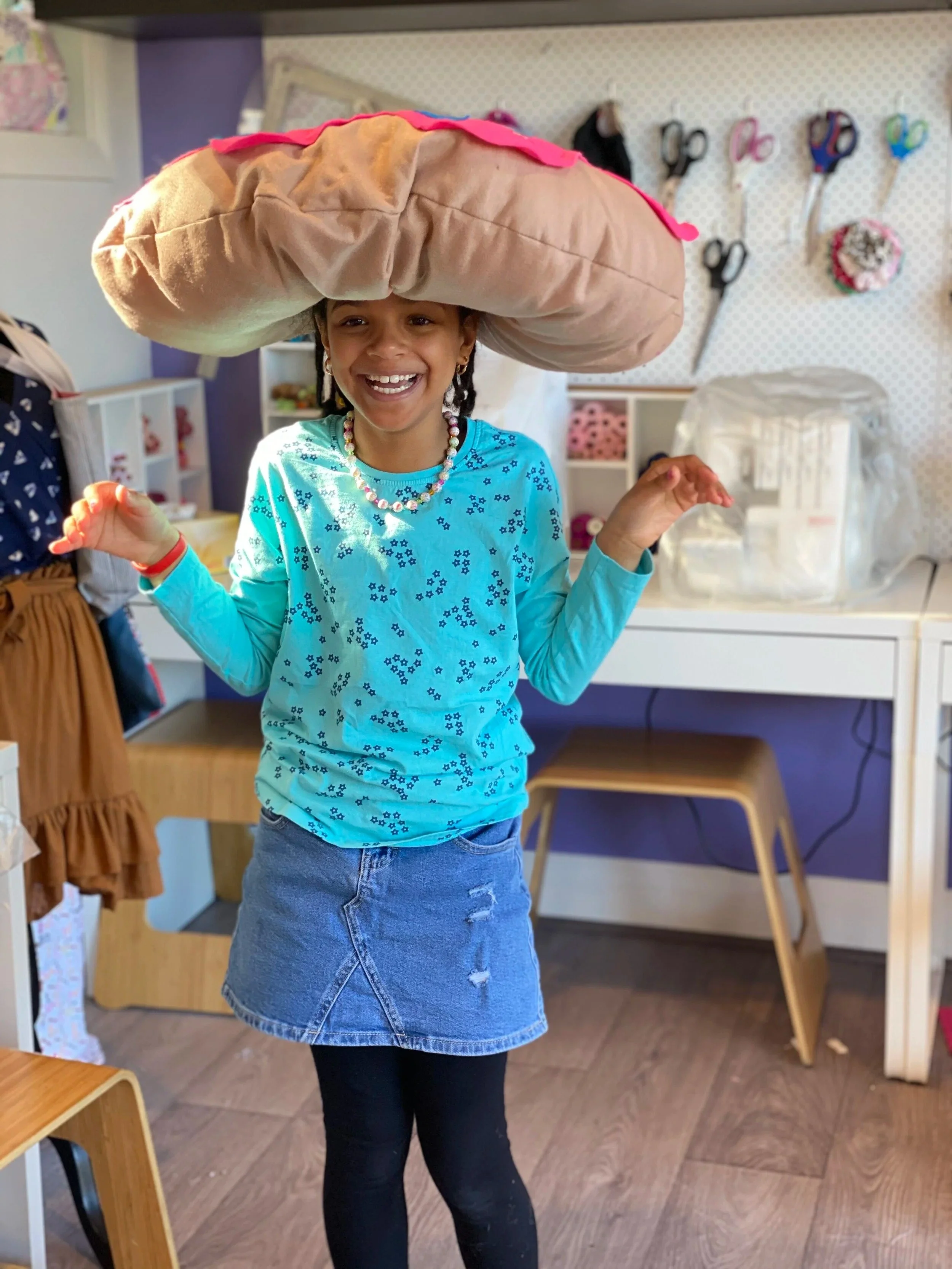 A girl smiling and laughing while wearing a large plush pillow on her head shaped like a hamburger bun in a craft room.