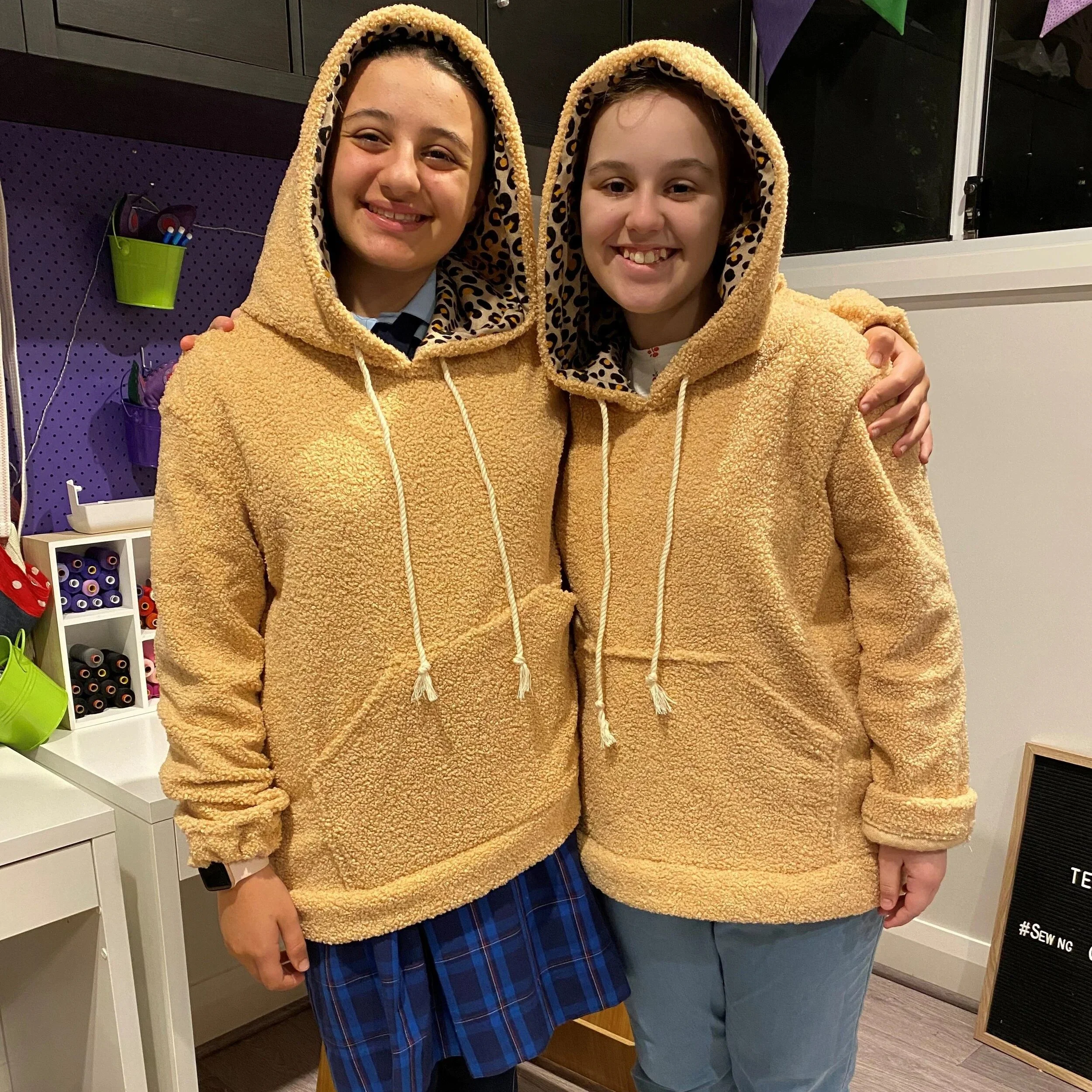 Two women standing close together, smiling, wearing matching beige fleece hoodies with leopard print lining inside the hoods, in a craft store.