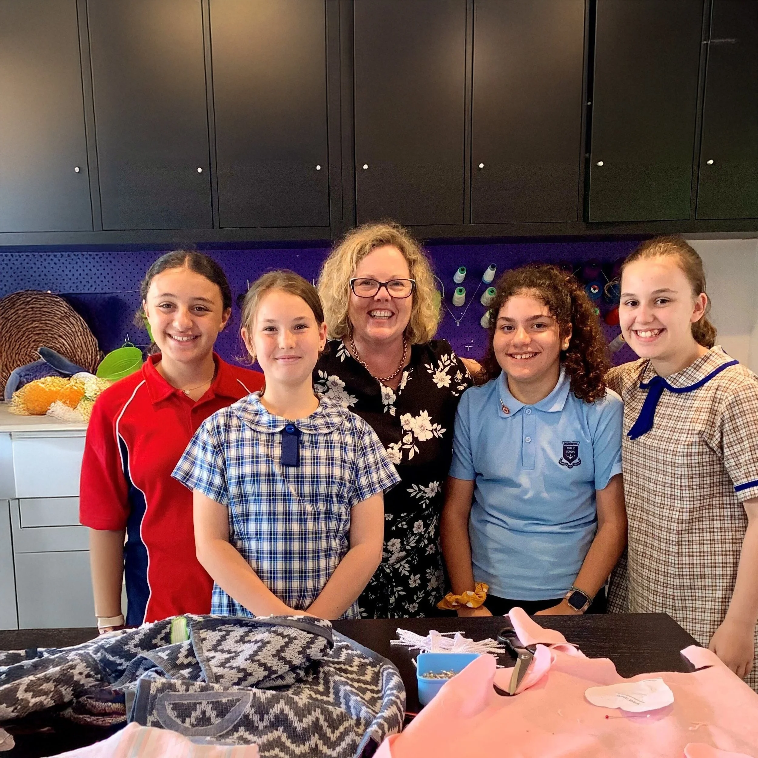 Group of five young girls and a woman standing together in a craft room, smiling at the camera, with sewing and craft supplies on the table in front of them.