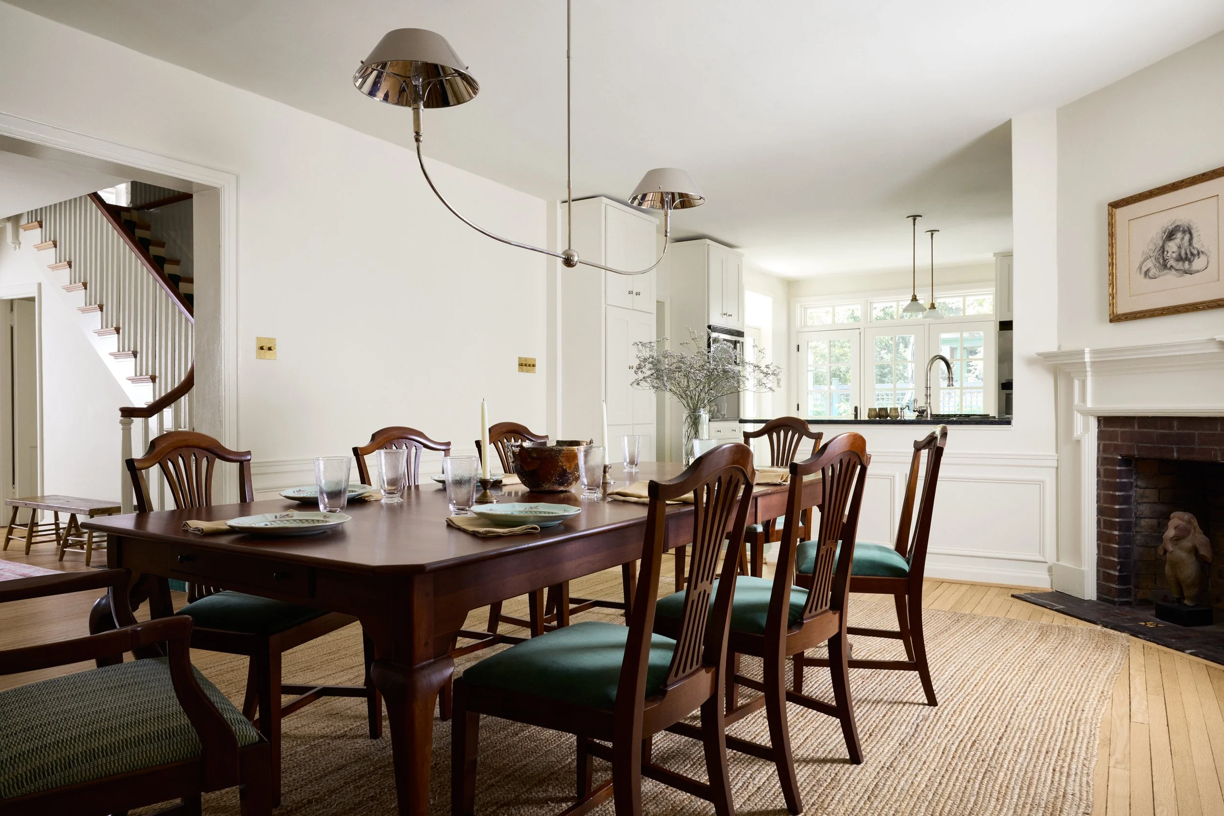 Dining room with a wooden table set for six, glassware, and candles. Kitchen in the background with large windows, fireplace on the right, and a framed sketch on the wall.
