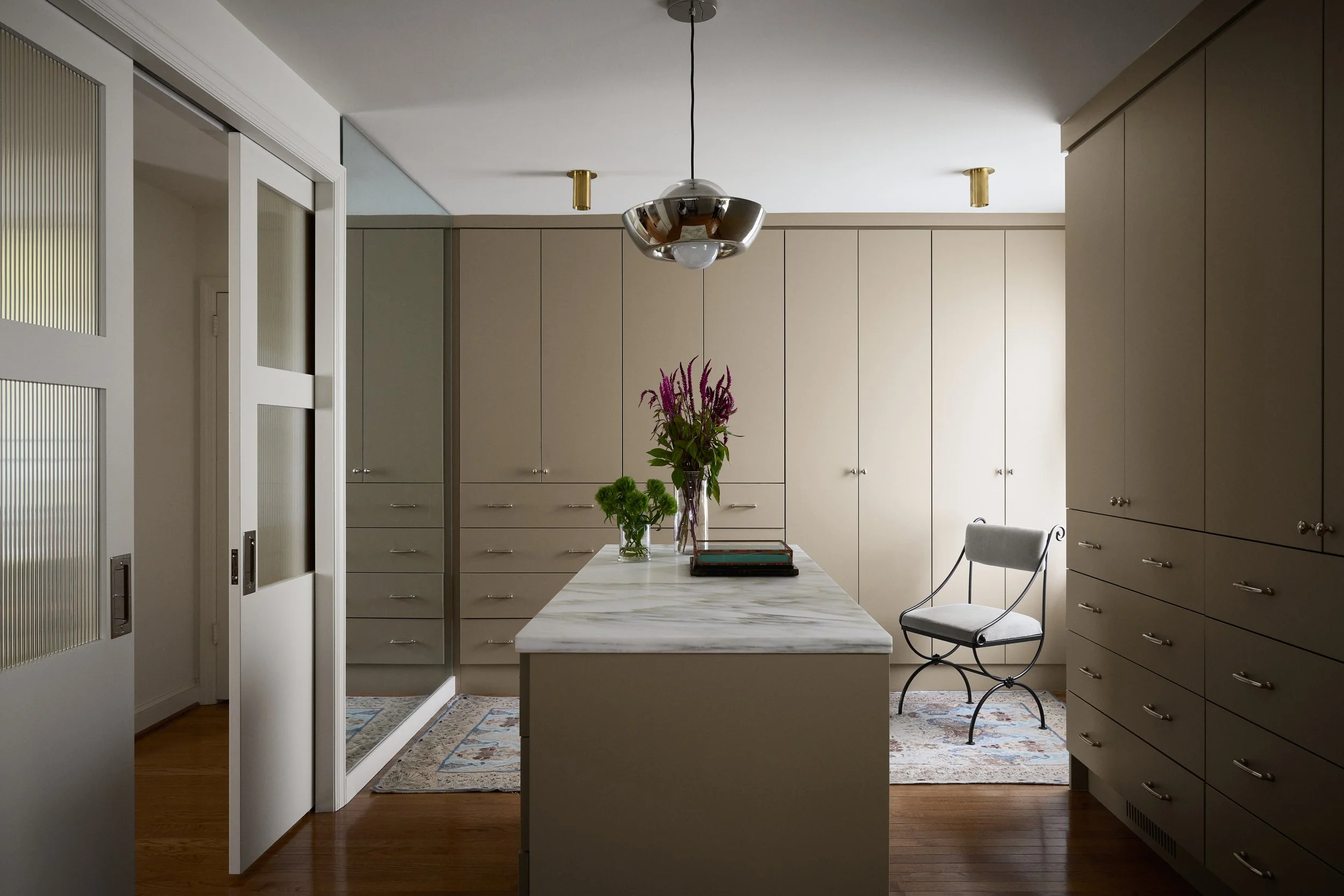 Kitchen with beige cabinets, a marble island with flowers, and a chair near a window with curtains.