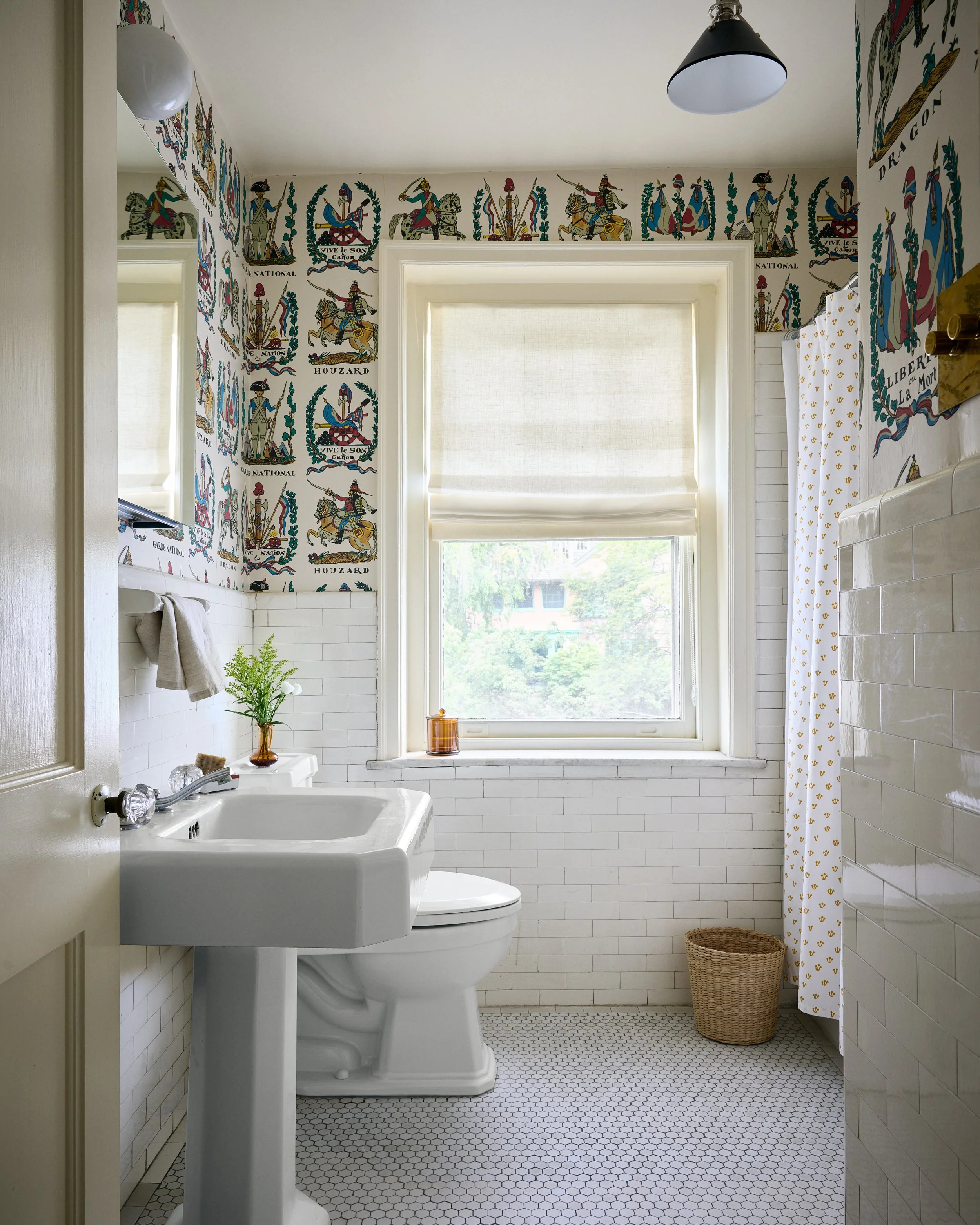 Bright bathroom with white subway tile walls, a vintage pedestal sink, a toilet, a large window with a beige roller shade, decorative wallpaper with medieval figures and banners, a small mirror, a towel rack with gray towels, a plant in a brown vase,
