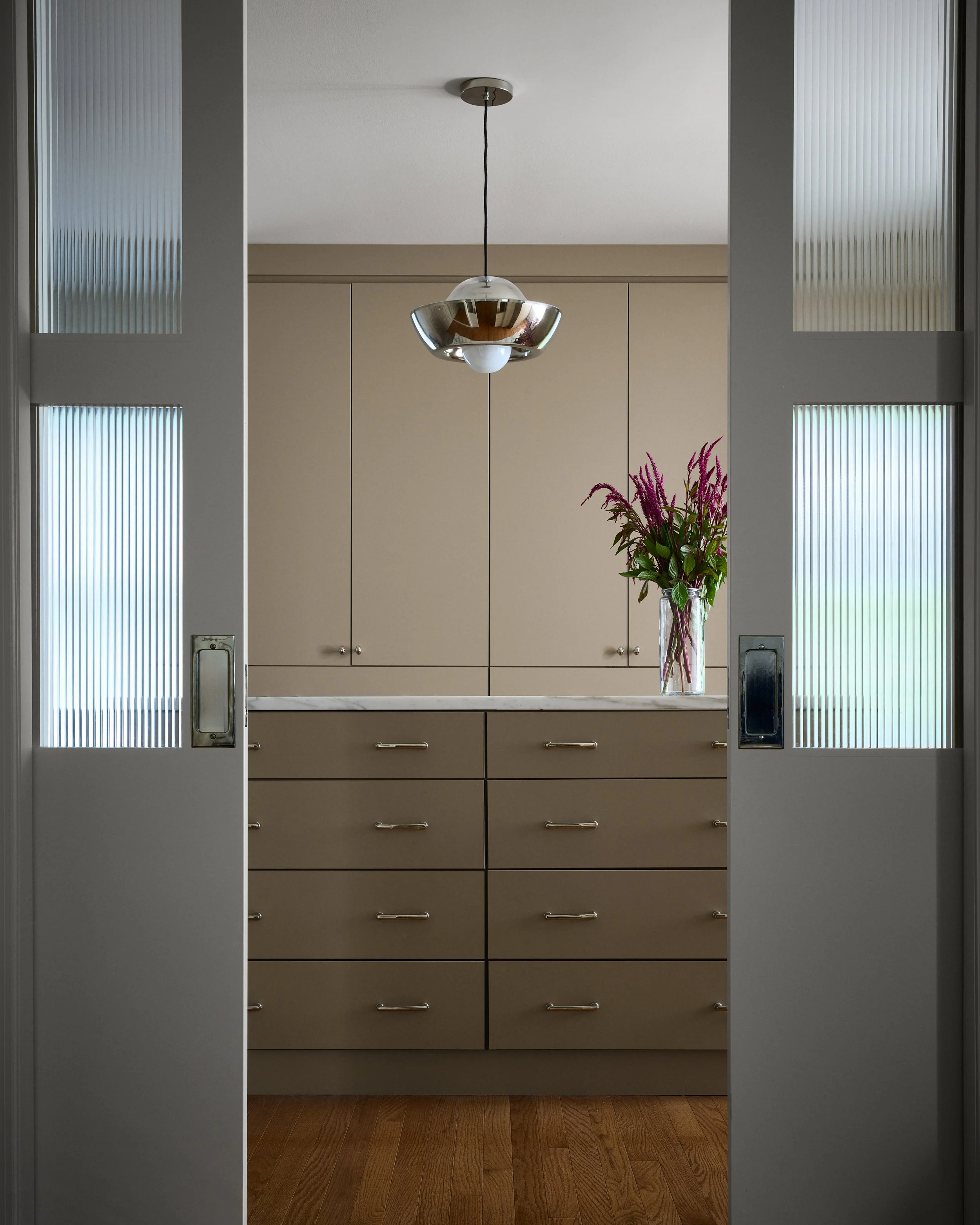 View through sliding glass doors into a modern kitchen with beige cabinets, a white countertop, a glass vase of pink and purple flowers, and a ceiling light fixture.