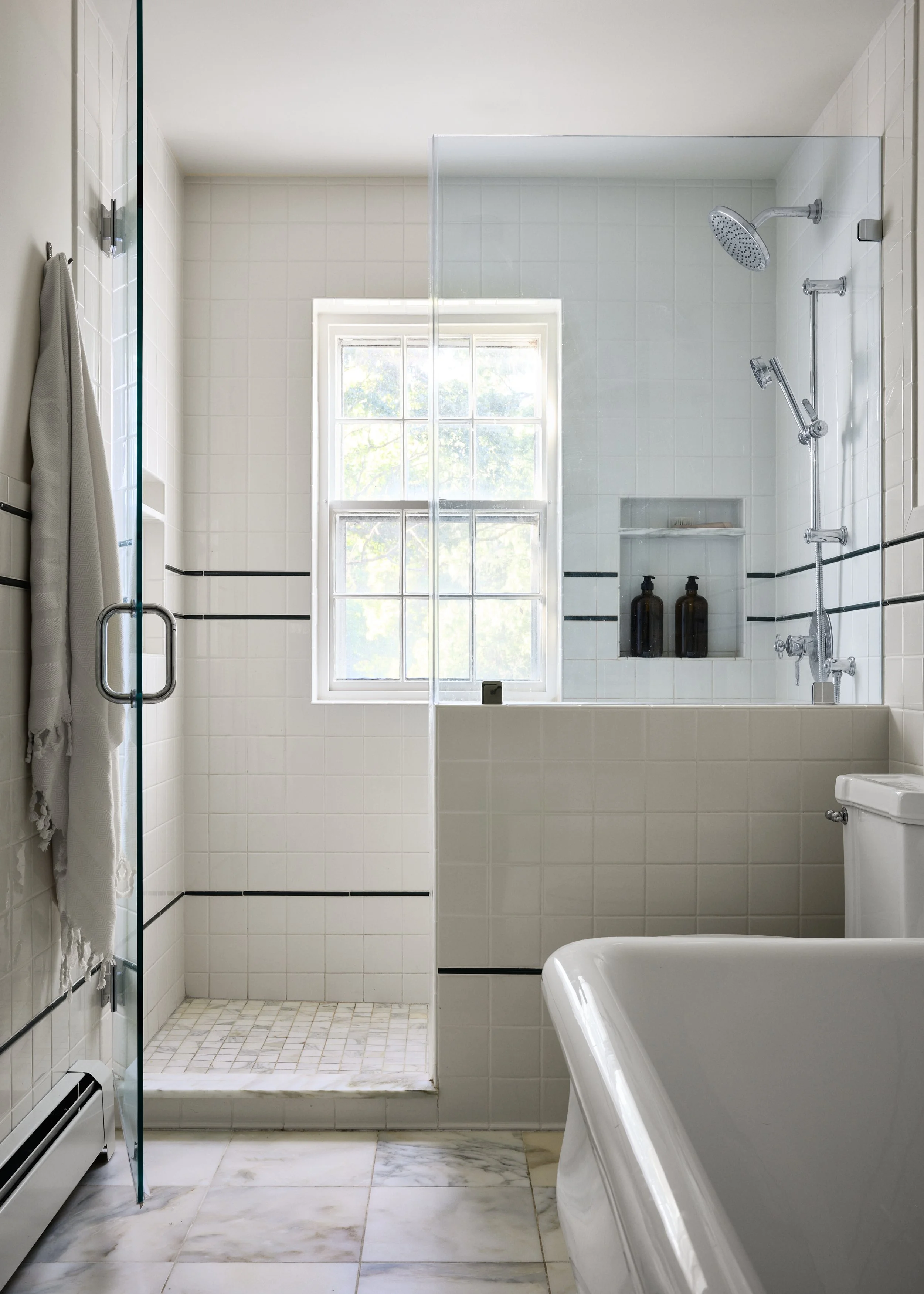 A bright bathroom featuring a walk-in shower with a glass door, a window, and a bathtub in the foreground. The shower area has white tiles with black accents and display bottles in a niche.
