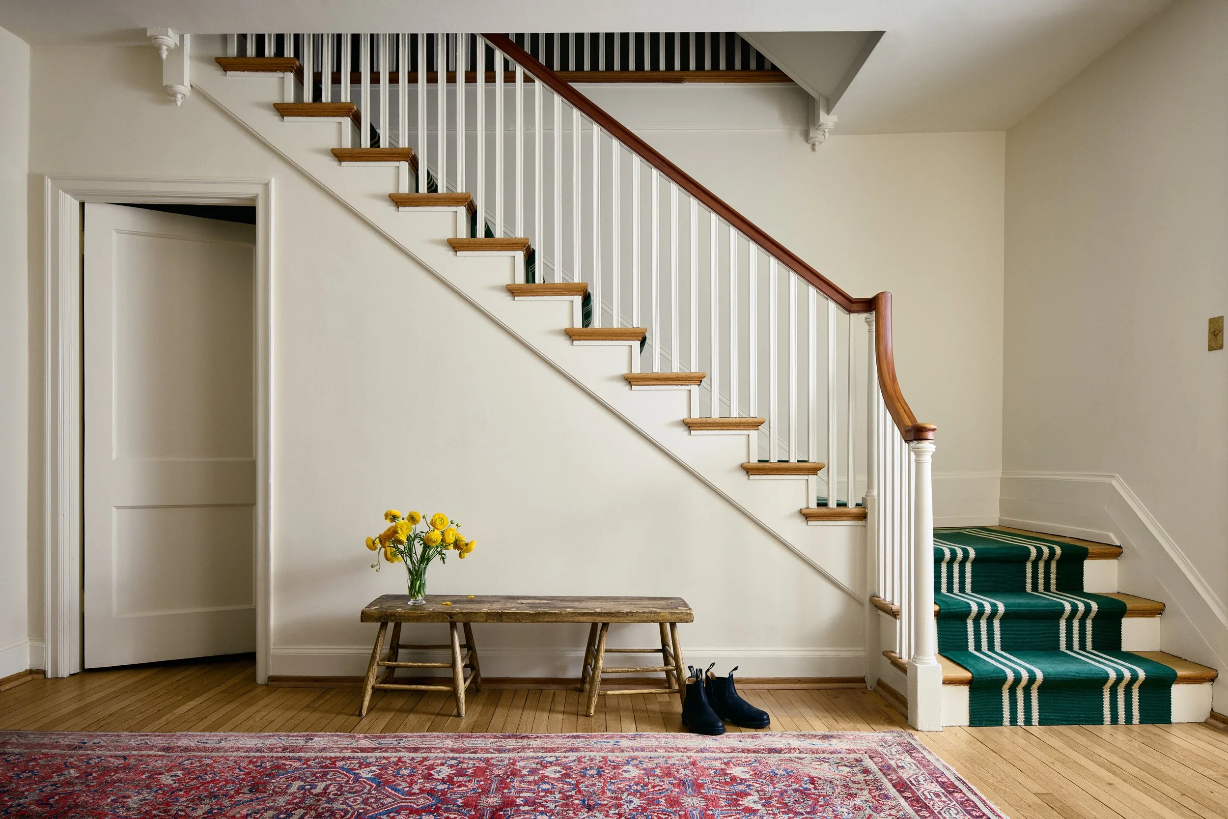 Interior view of a house entryway with a staircase and a small wooden bench with a vase of yellow flowers, a pair of black boots on the floor, and a patterned rug.