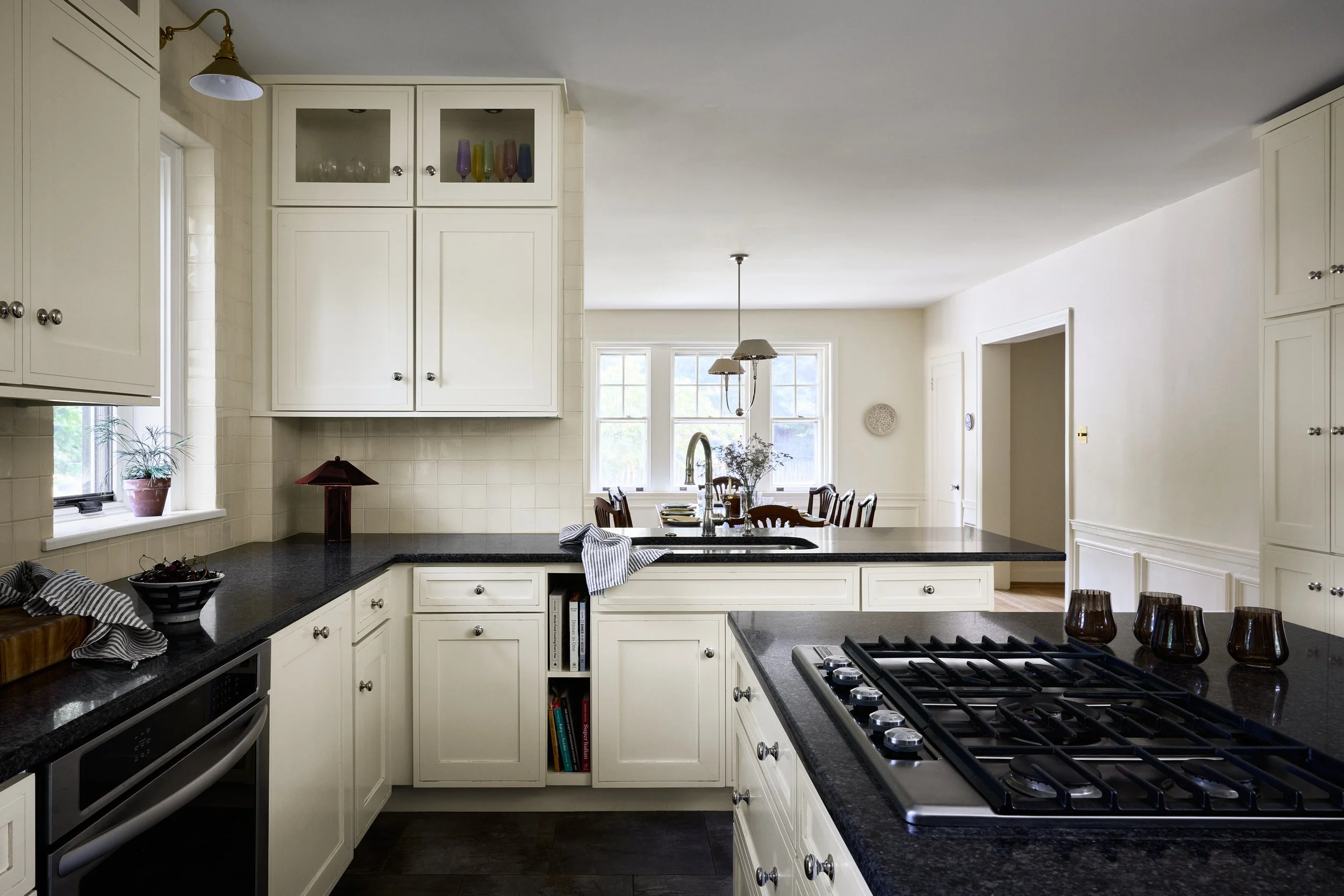 Bright white kitchen with black countertops, a central island with a stove, and a dining area with a table and chairs in the background.