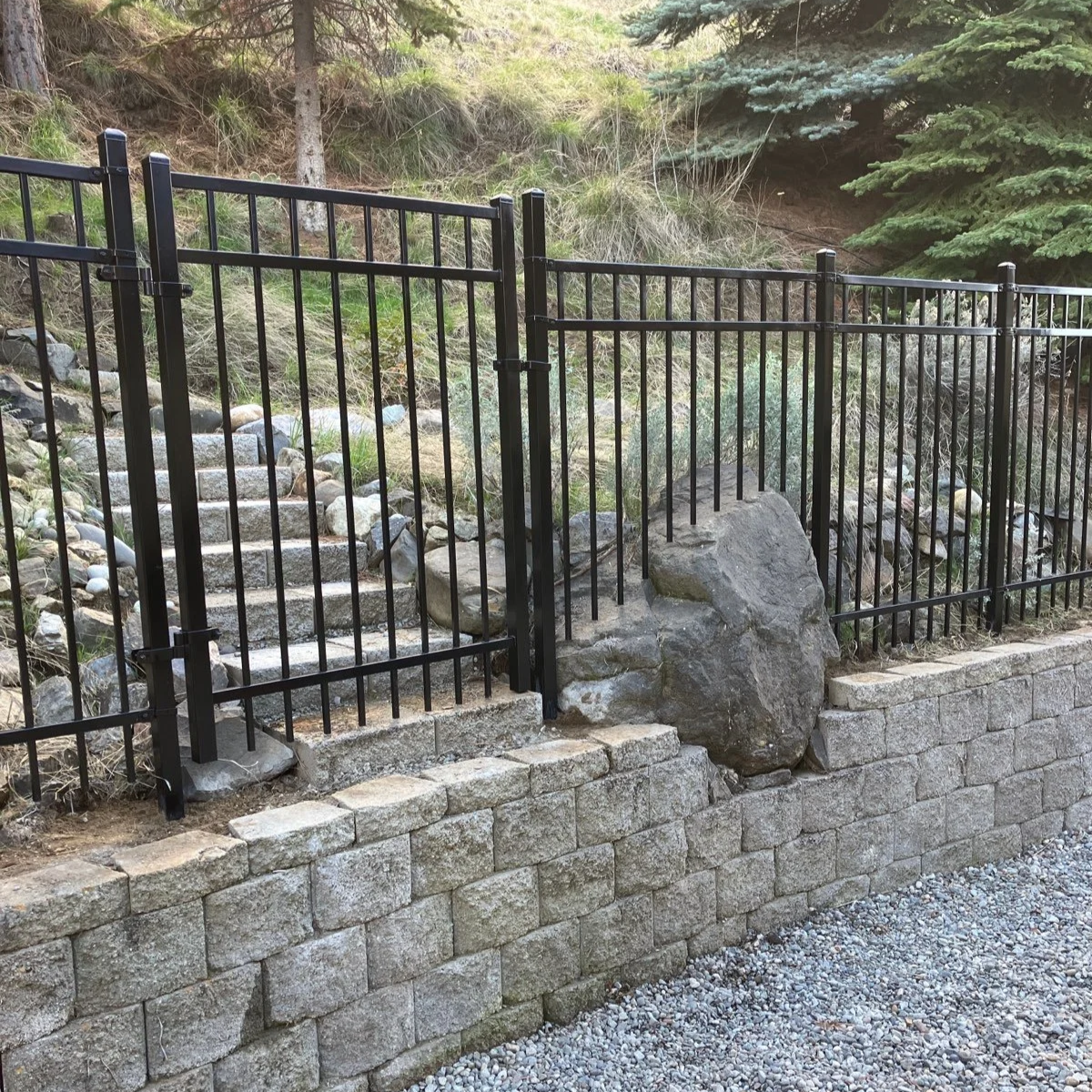 A black metal fence surrounds a stepped stone pathway leading up a hillside with rocks and greenery, including pine trees.