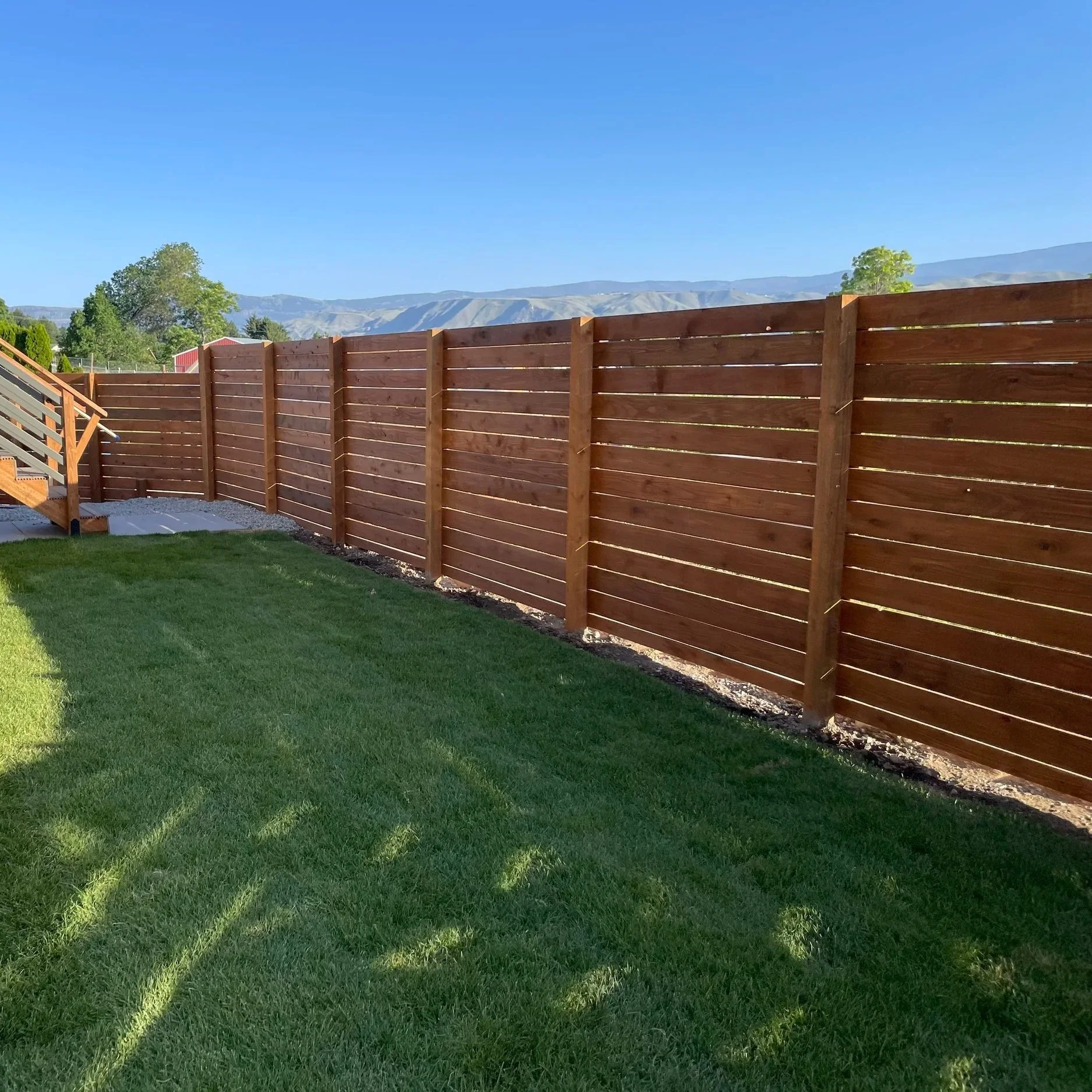Backyard with a wooden fence, green grass, trees, and distant mountains under a clear blue sky.