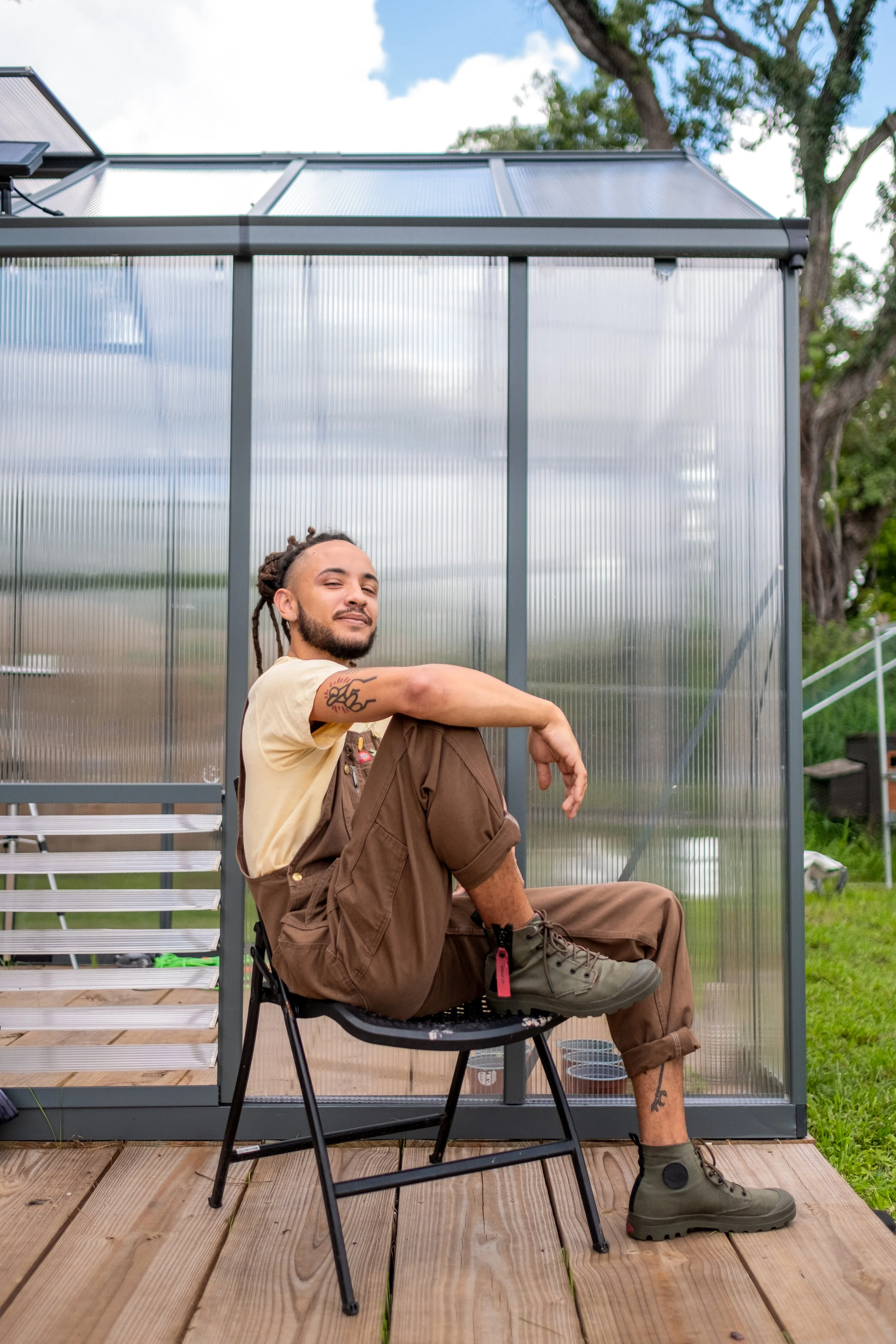 A grinning man sitting in a folding chair in front of a greenhouse with his arm up on his knee