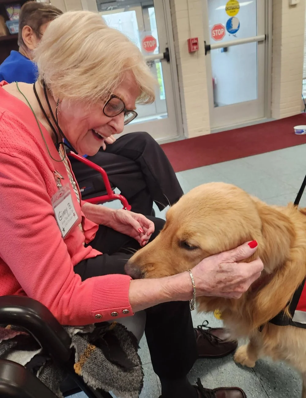 An elderly woman wearing glasses, a pink sweater, and a name tag with 'Gina' on it, is smiling and holding the face of a golden retriever dog. She is sitting in a wheelchair. The dog is standing and looking at the woman, with its head gently held in her hands. There are two people seated behind them near a glass door with stop signs.