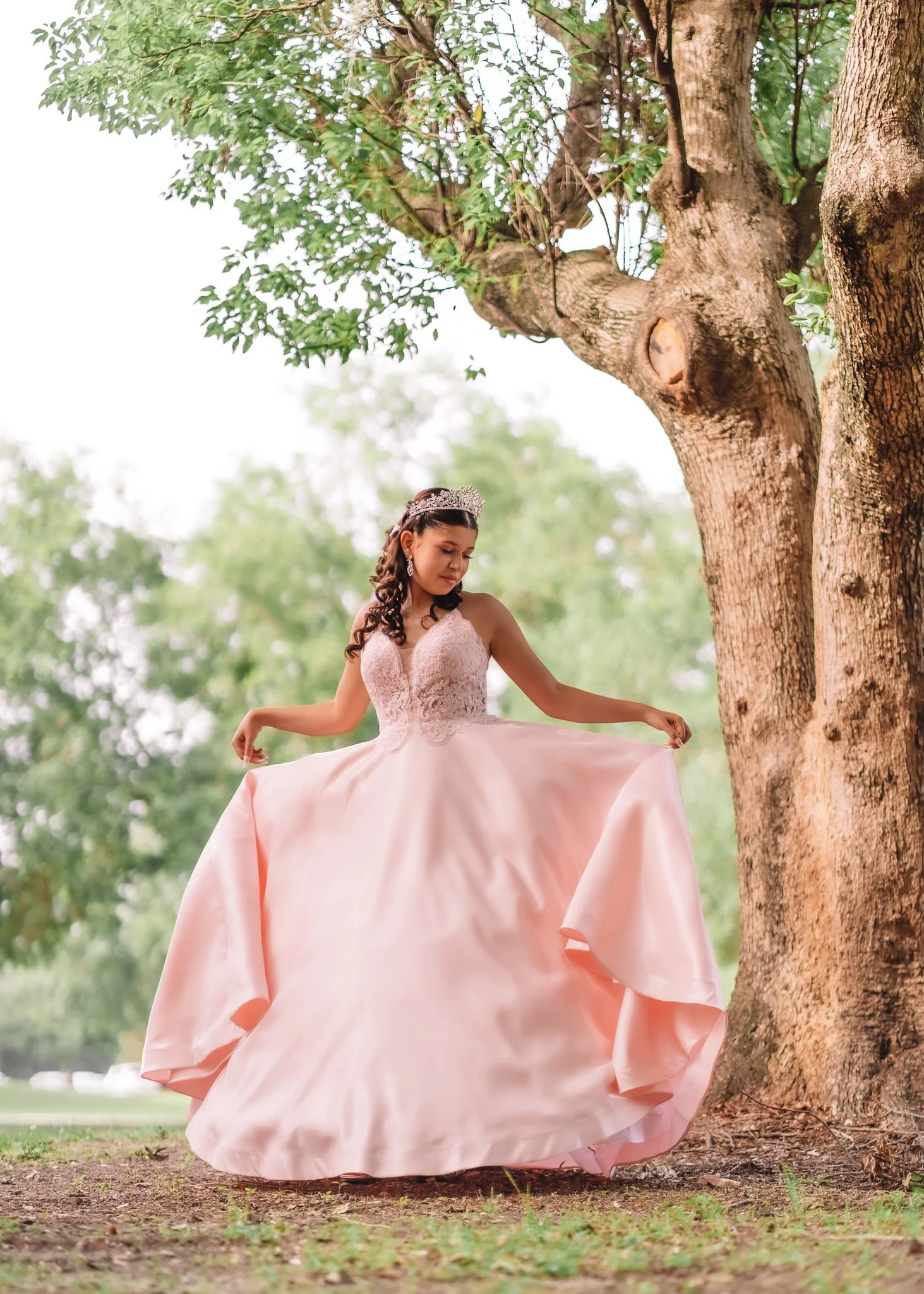 A young woman in a pink ball gown with lace bodice, holding the sides of her wide skirt while standing outdoors near a large tree, with green leaves and blurred background.