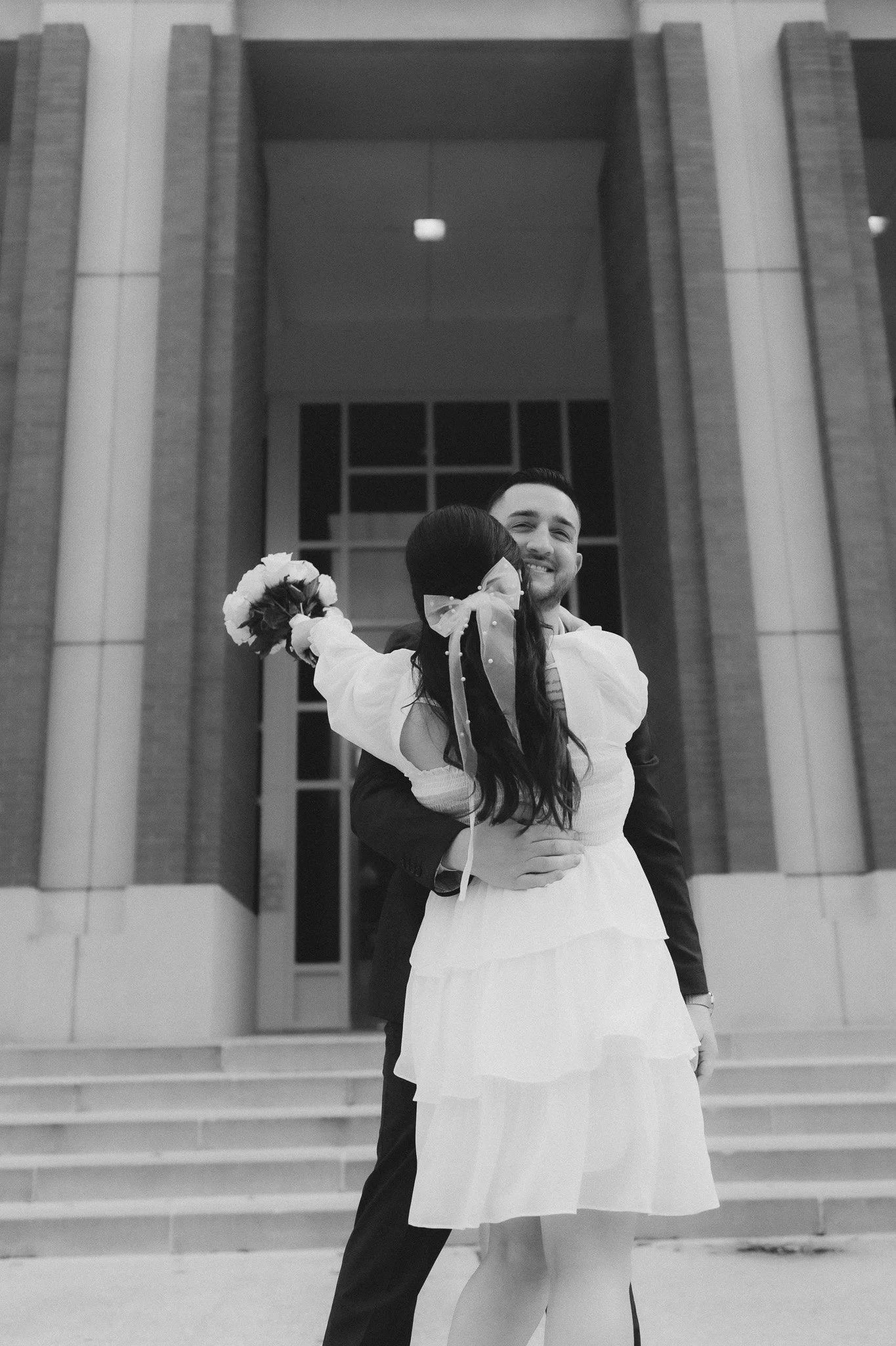 A man and woman embrace in front of a building with large glass windows, the man smiling and the woman holding a bouquet of flowers.