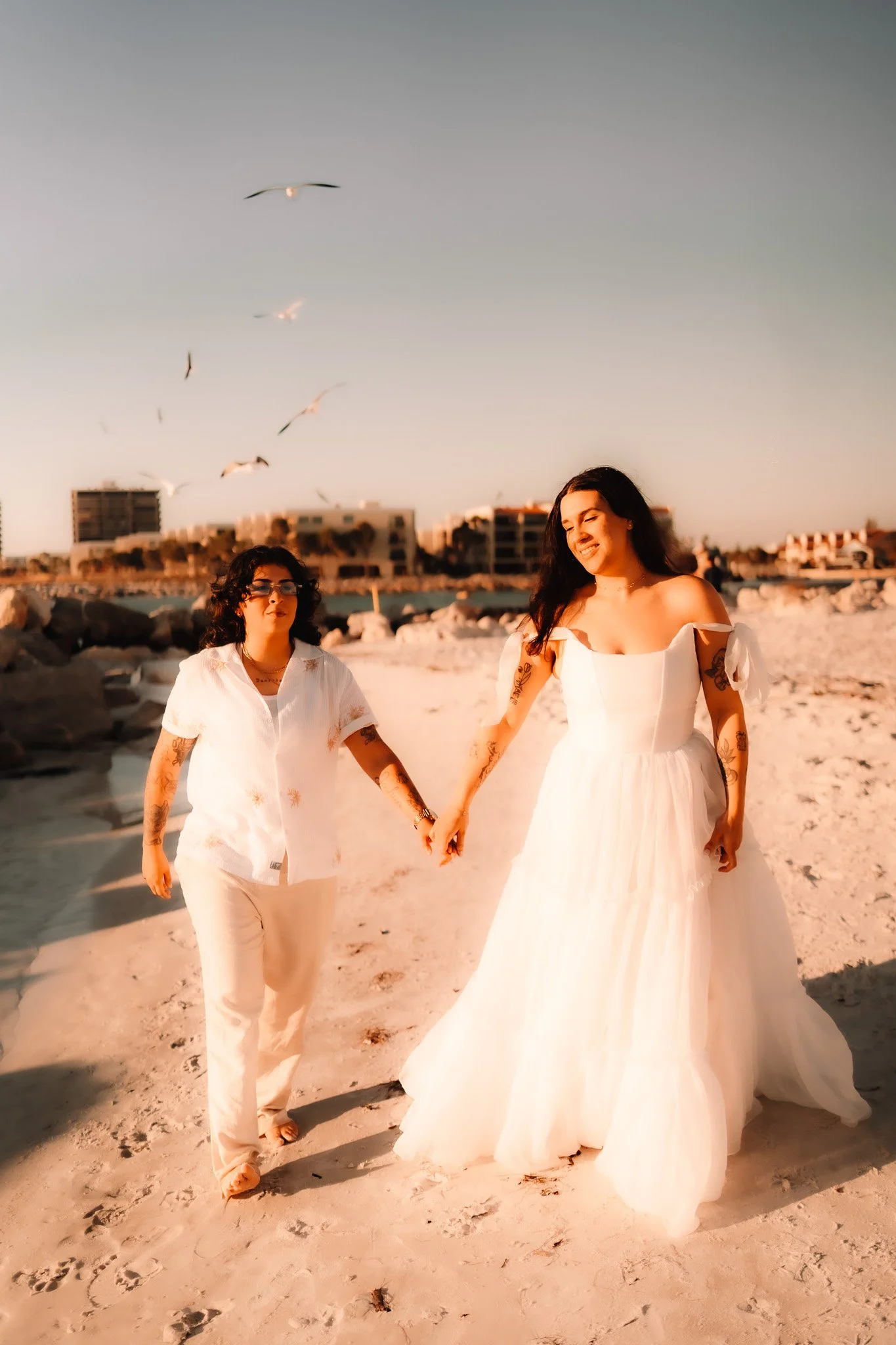 Two women walking hand in hand on the beach during sunset, one in a white wedding dress and the other in a white outfit with tattoos, seagulls flying overhead, buildings in the background.