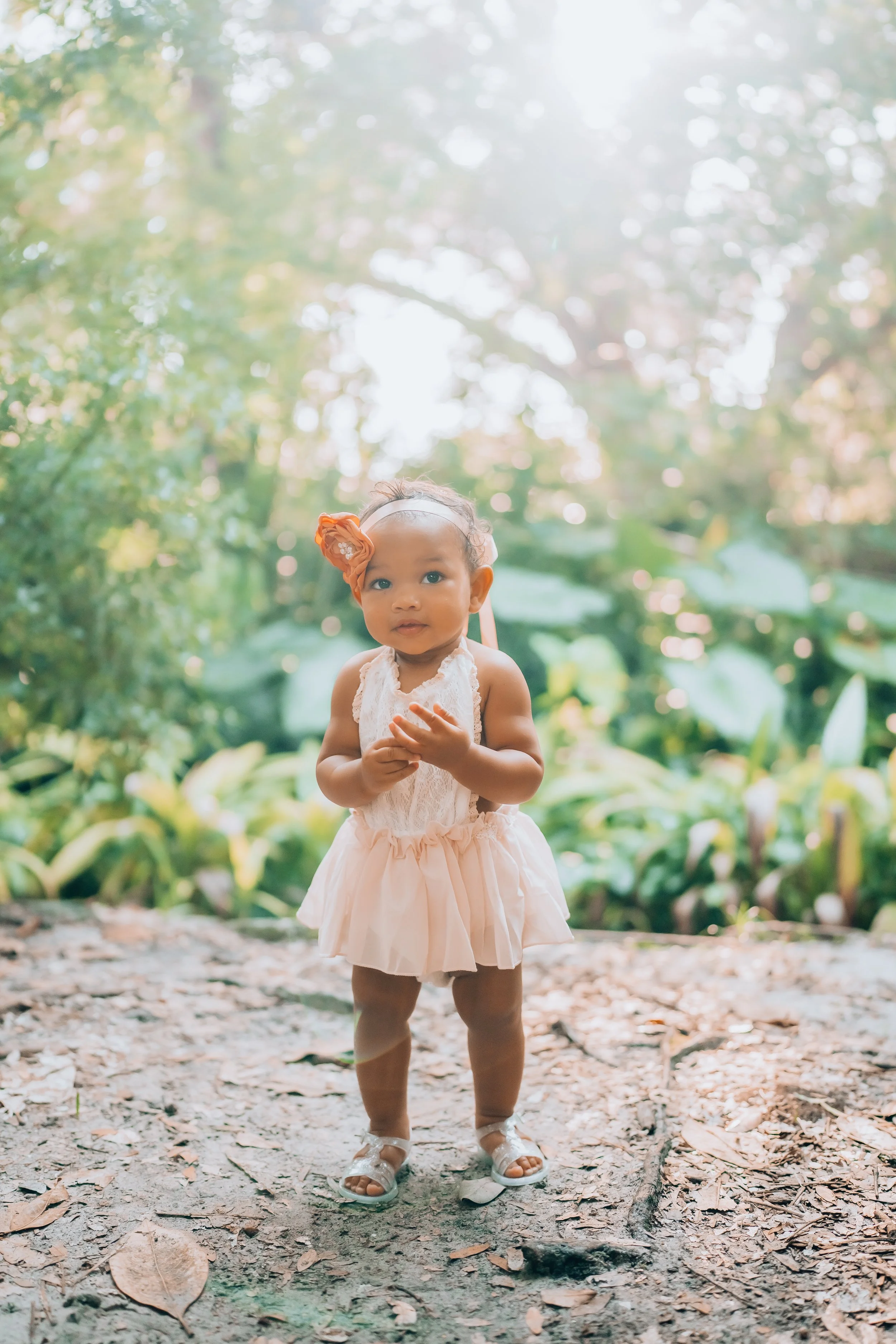 A young girl standing outdoors on a trail, surrounded by green foliage, wearing a light pink dress, sandals, and a floral headband.
