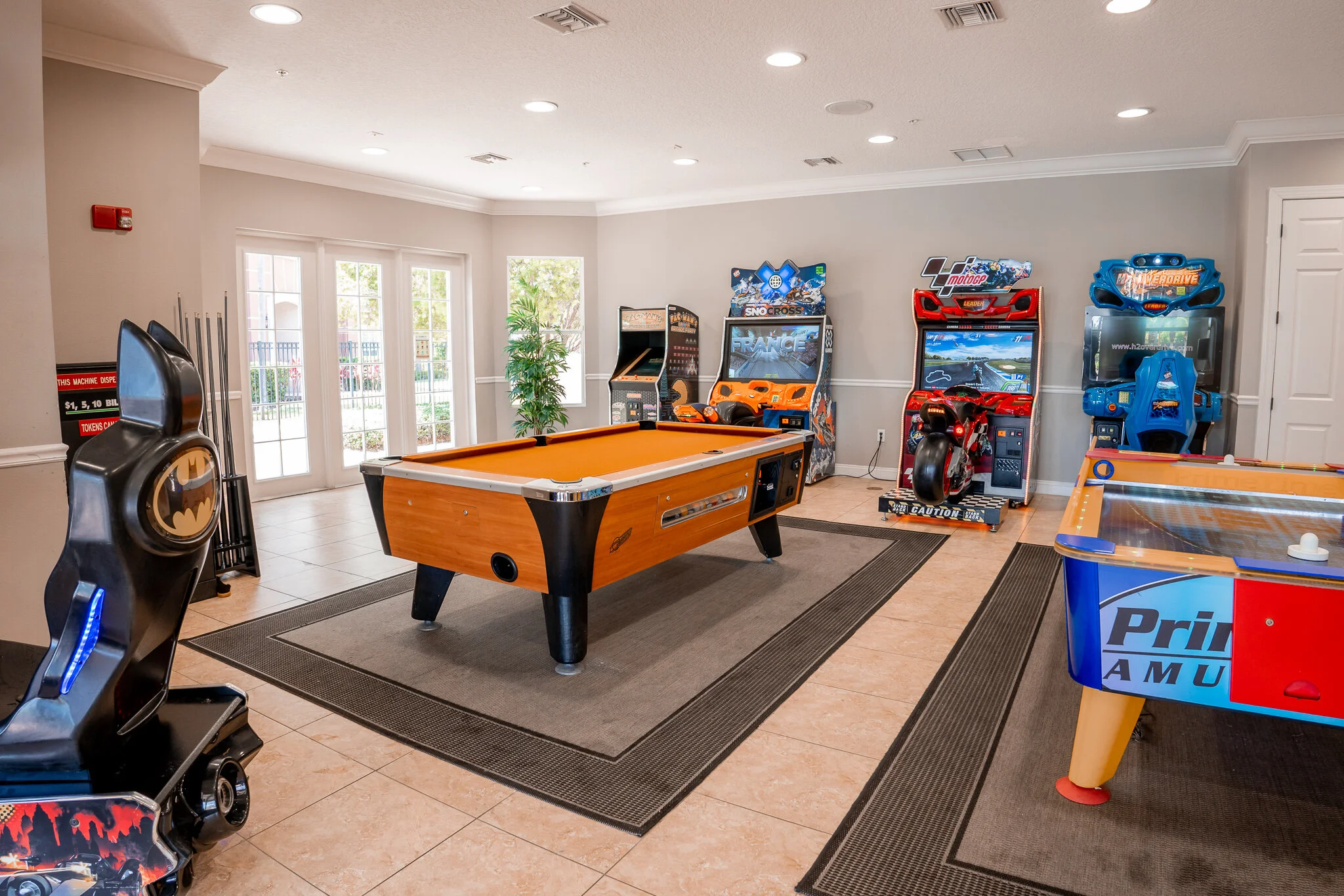 Indoor arcade room with a pool table, multiple arcade racing games, an air hockey table, Batman-themed arcade game, and doors leading outside.