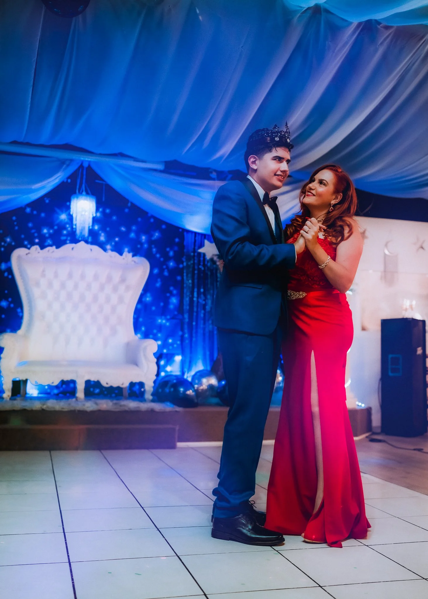 A young man and woman dance together at a formal event. The man is wearing a black tuxedo with a bow tie and a crown, and the woman is wearing a long red gown with a slit and earrings. They are holding hands and smiling at each other, with a decorate
